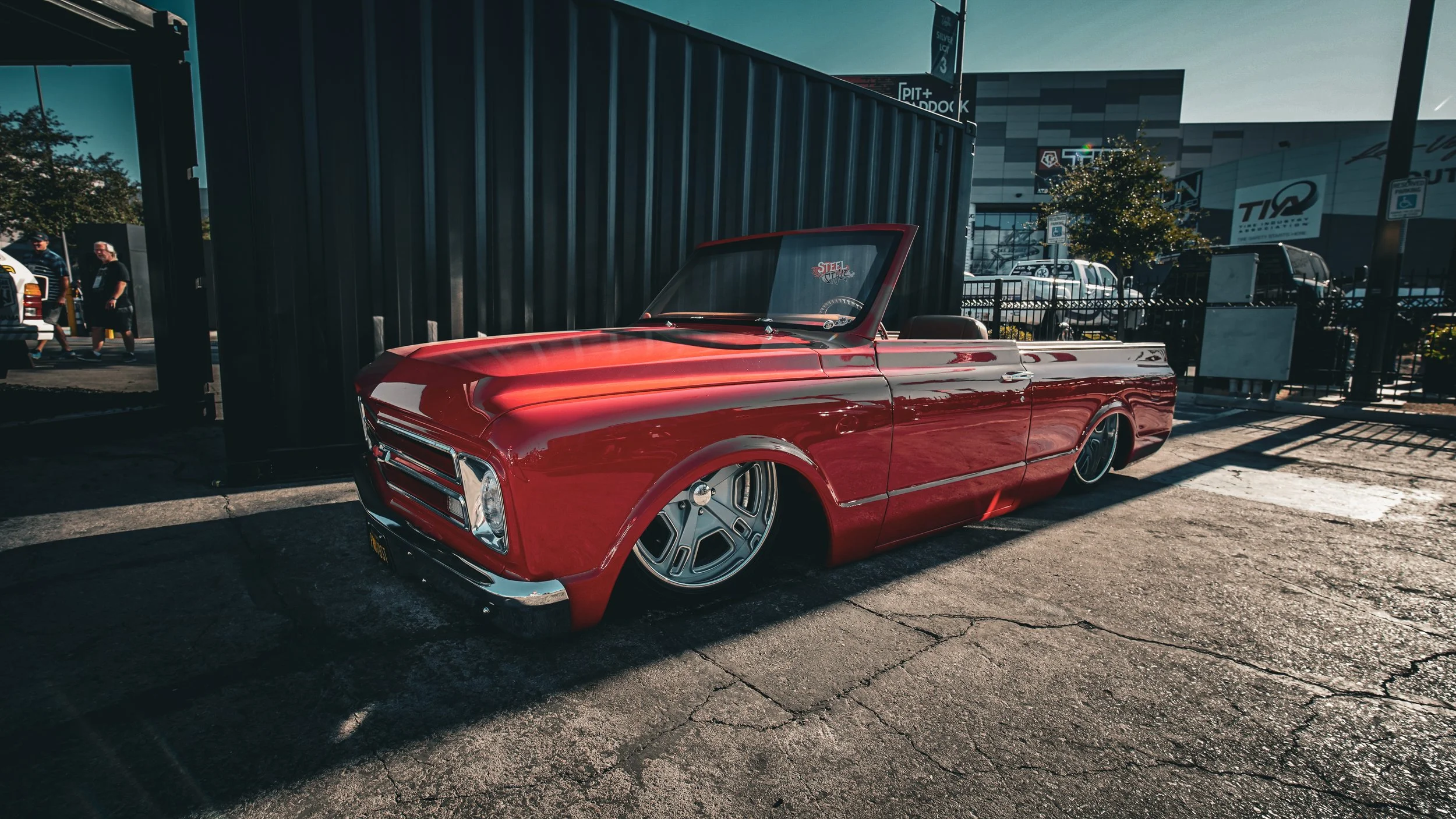 Red vintage convertible car parked on asphalt outside in a commercial area, with people and buildings in the background, and a tree decorated with lights.