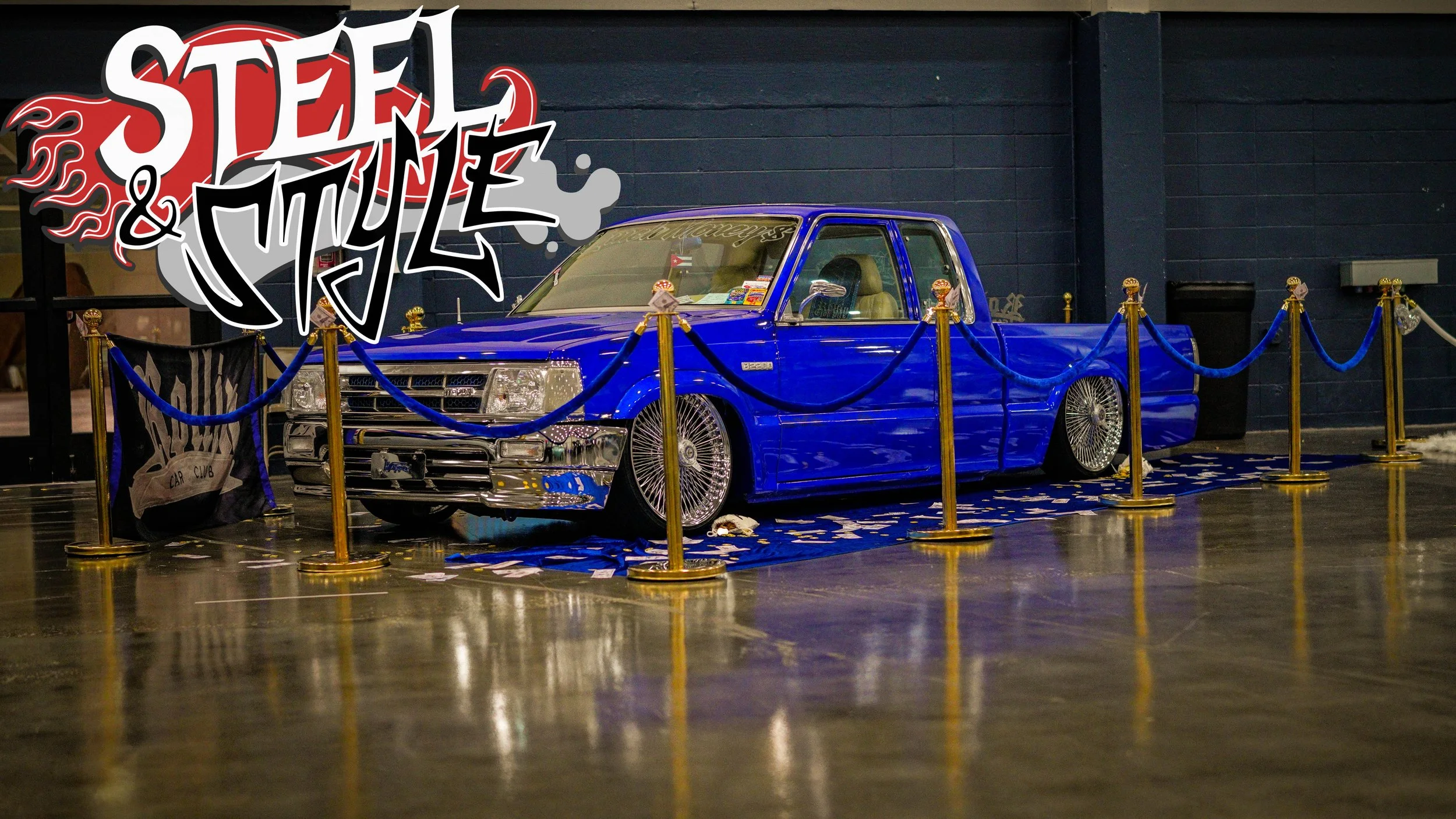 A blue lowrider truck with chrome details is displayed inside a roped-off area at an indoor event, with a large 'Steel & Style' banner above it.