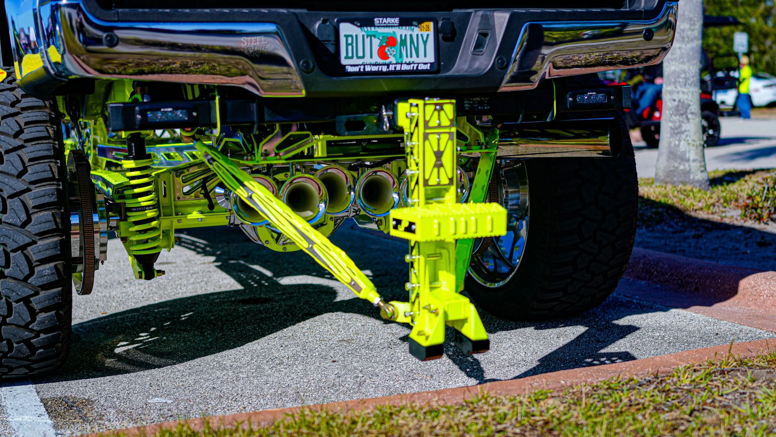 Close-up of a lifted pickup truck with custom bright yellow suspension and exhaust system, displaying large tires and vehicle licensing plate that reads 'BUT ON MY'.