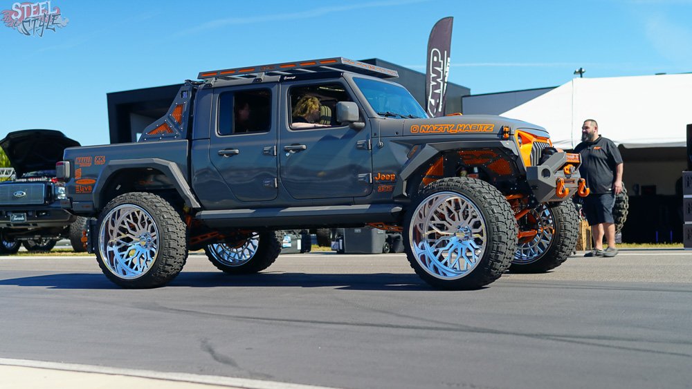 A modified Jeep truck with oversized, intricate alloy wheels and off-road tires, displayed at an outdoor car show on a sunny day. The vehicle has custom orange accents and branding, with people walking around in the background.