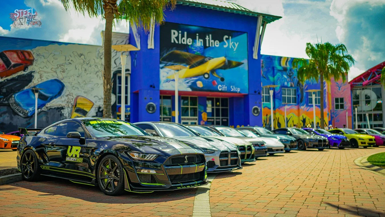 Black Ford Mustang Shelby GT500 leading a lineup of performance cars outside Dezerland Park in Orlando during the Velocity Rally, with the Ride in the Sky attraction in the background.