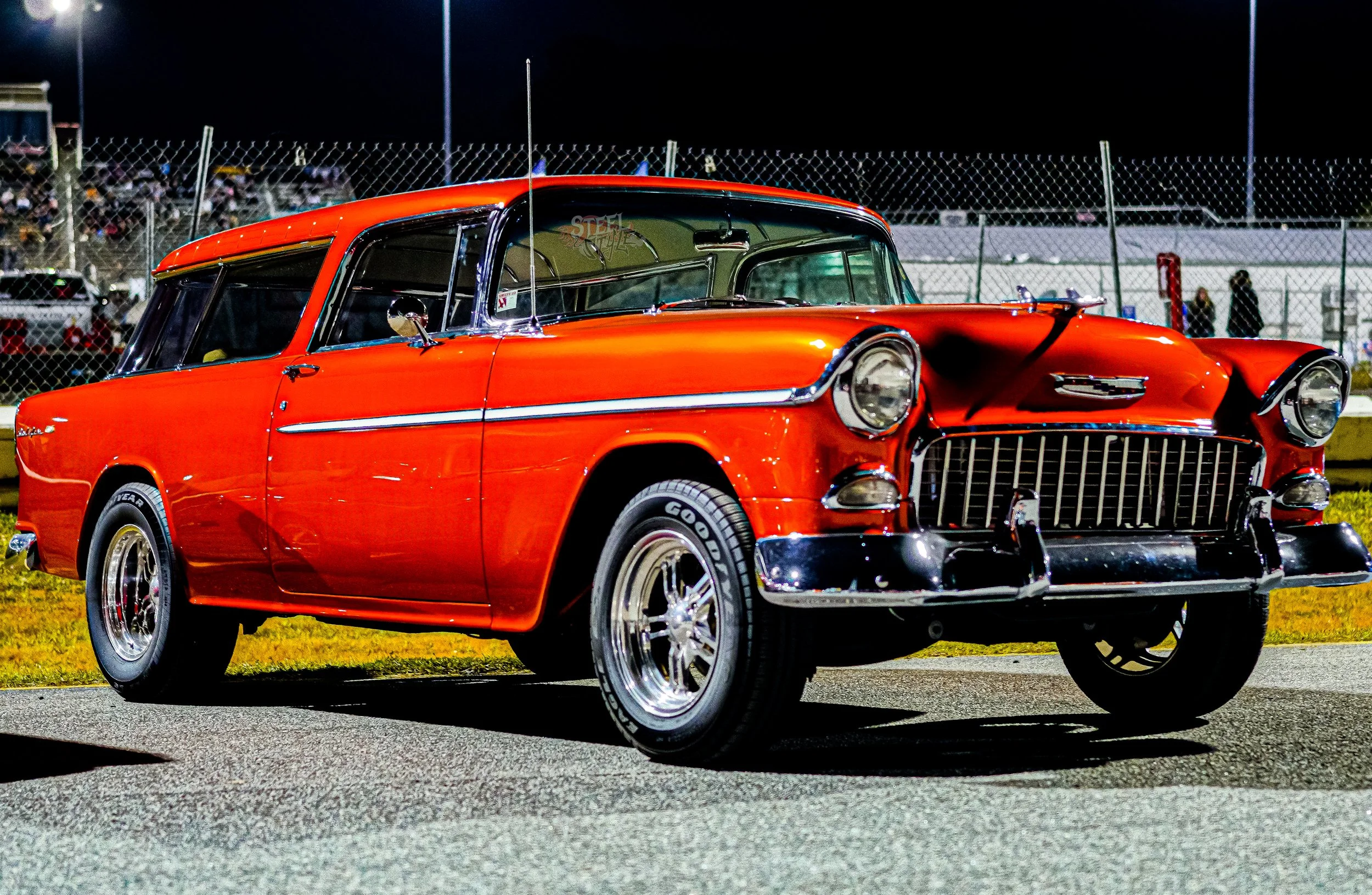 A vintage red car parked on a racetrack at night with a chain-link fence and spectators in the background.