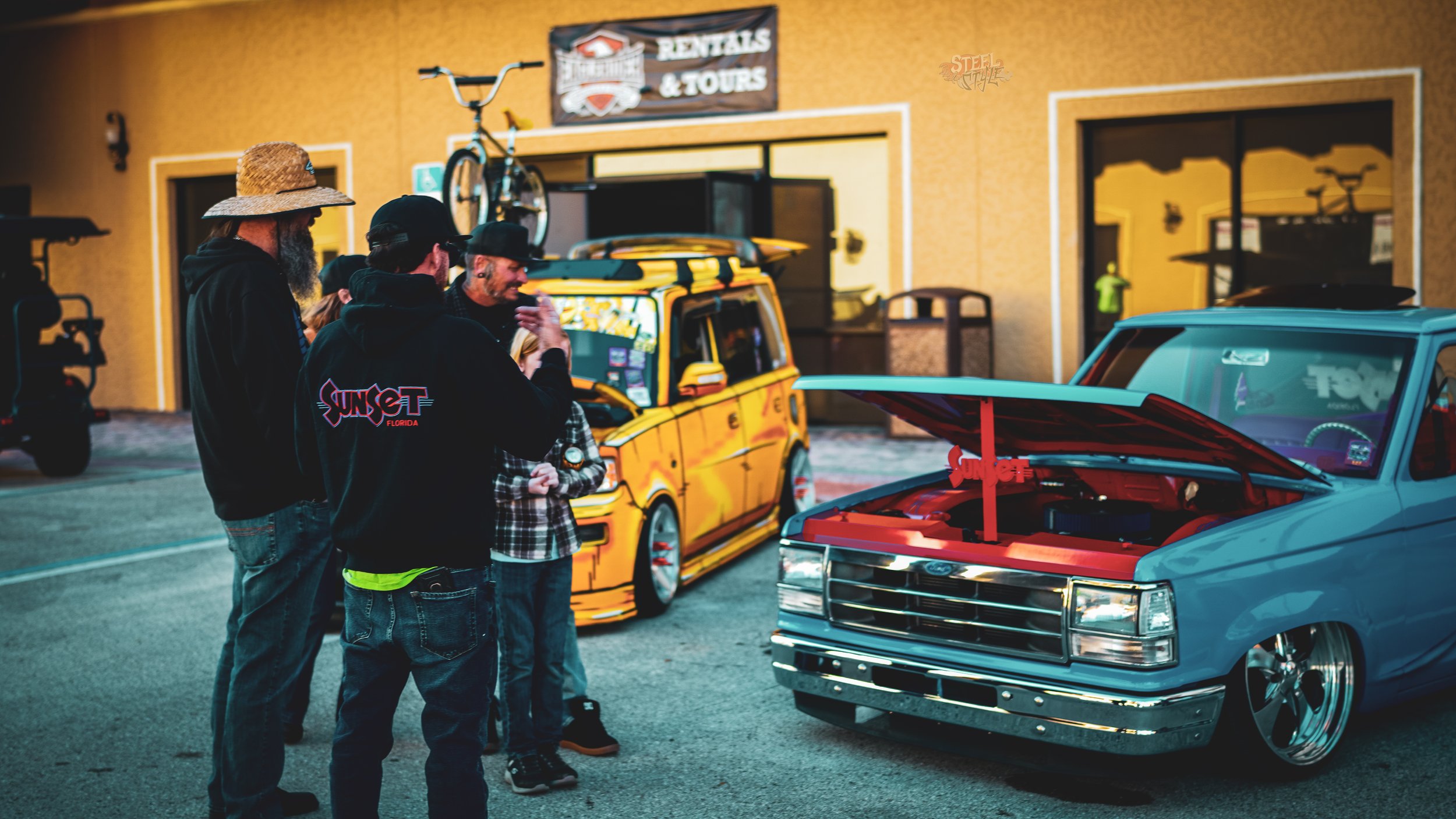 A group of people gathered around a blue vintage car with its hood open, parked outside a building with a sign for rentals and tours. Other vehicles and bicycles are visible in the background.