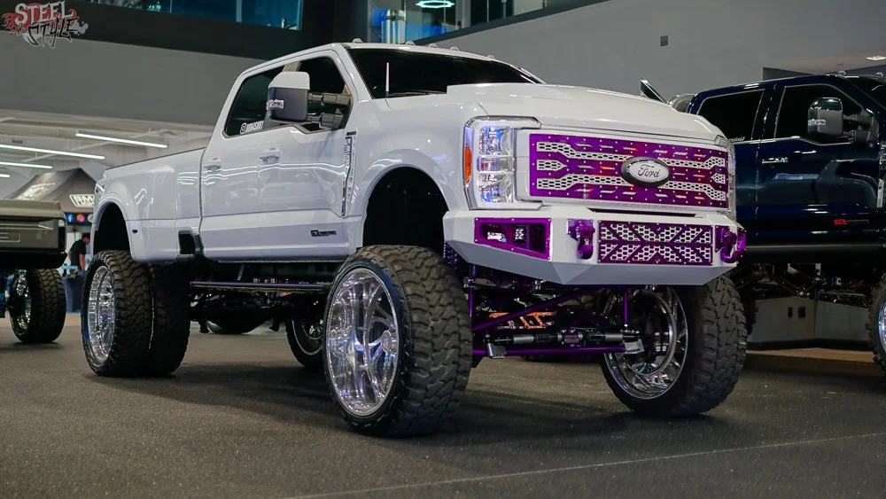 A customized white Ford pickup truck with large off-road tires, chrome rims, and purple accents on the front grill and bumper, displayed at an auto show.