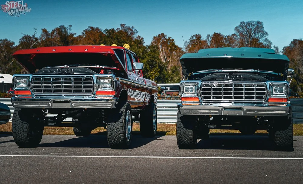 Two lifted classic pickup trucks with hoods open, parked side by side on a racetrack, with trees and blue sky in the background.