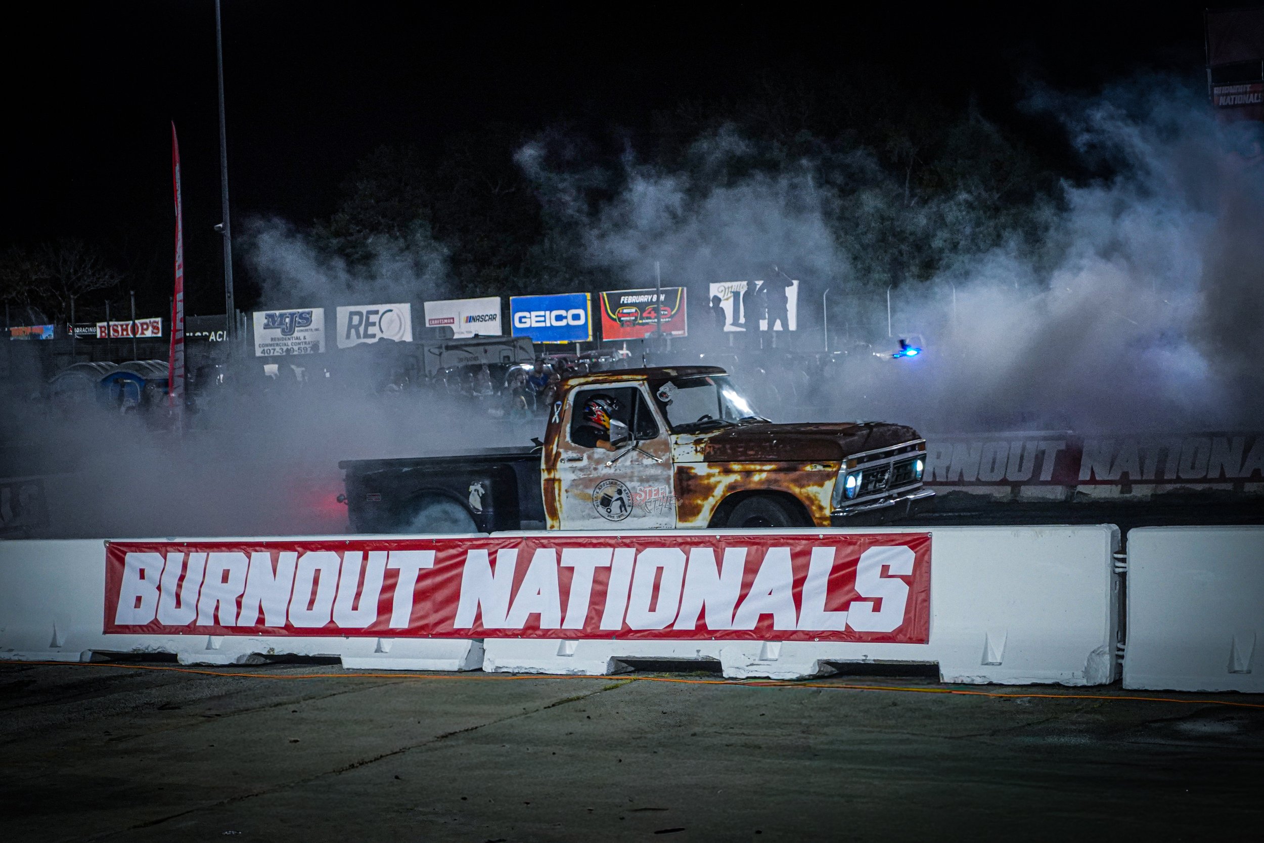 A rusty pickup truck on a racetrack at night during burnout Nationals, billowing smoke with banners and spectators in the background.