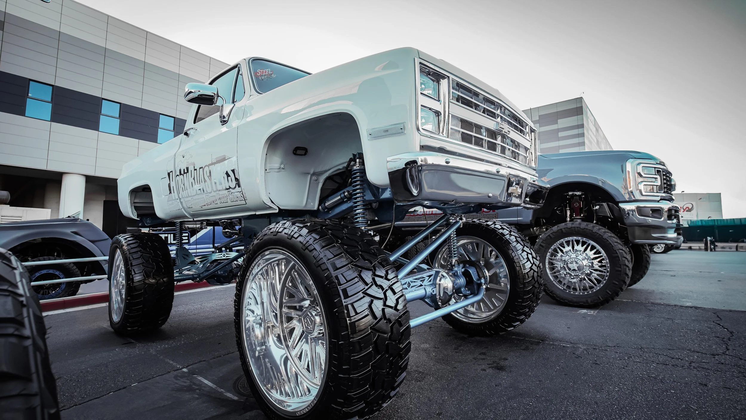 A custom lifted truck with large off-road tires and chrome wheels parked outdoors near a modern building.