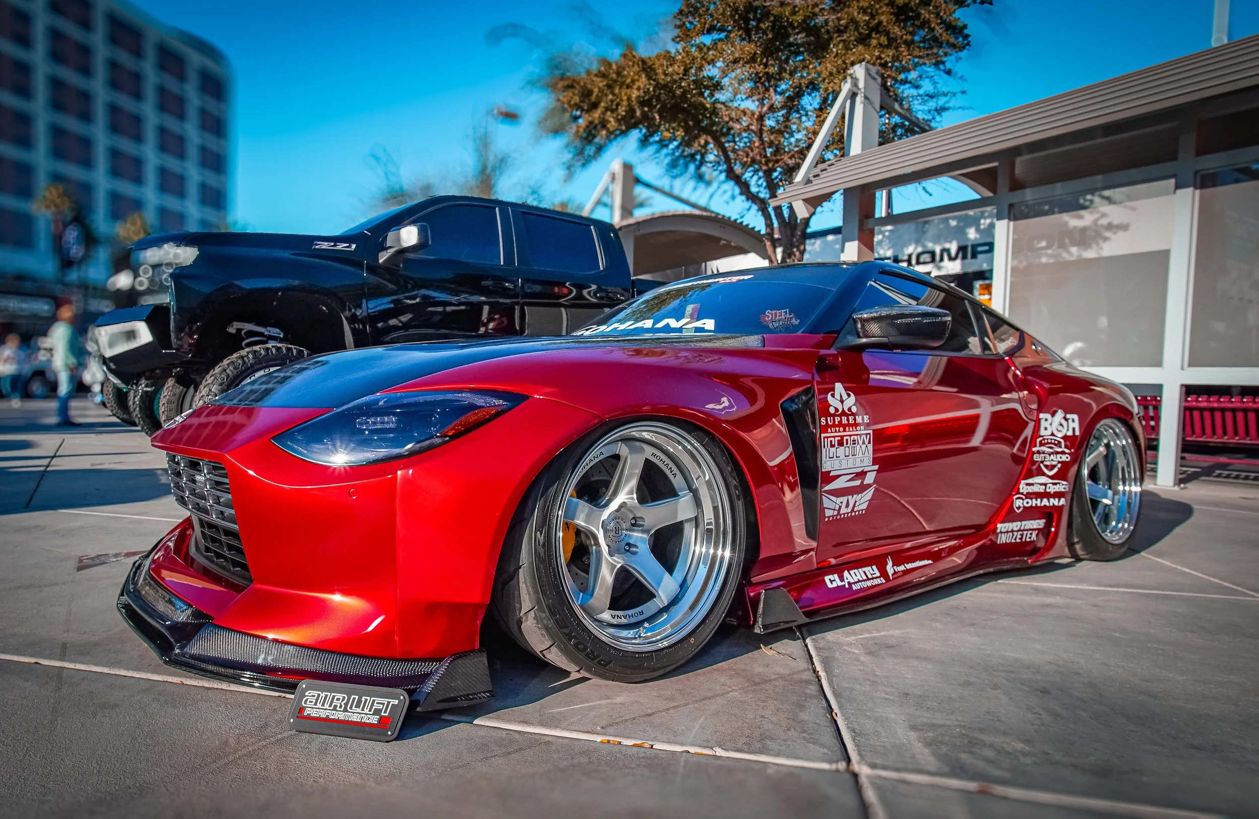 Red and black racing car with sponsor decals parked next to a black pickup truck on a city street during daytime.