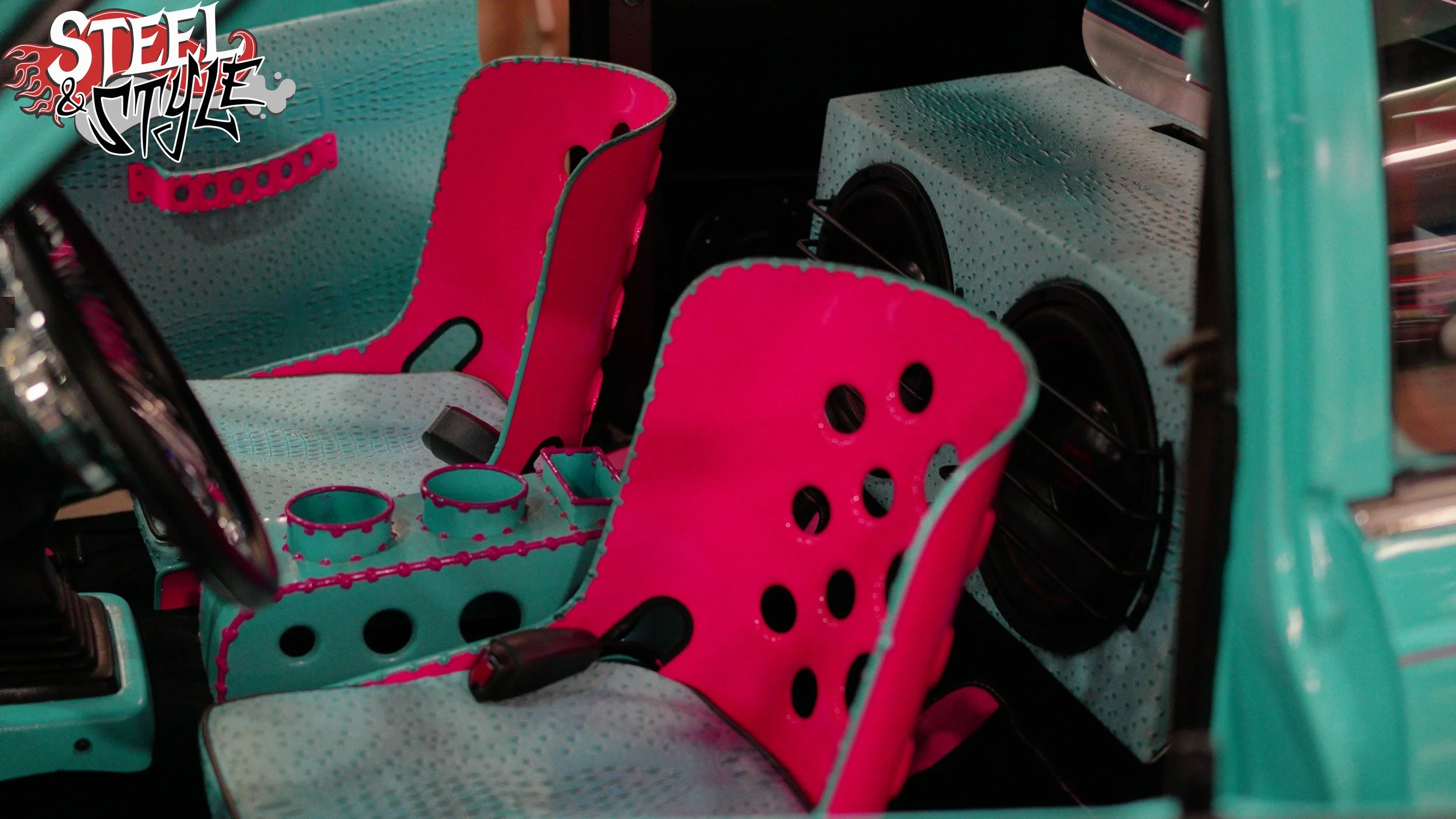 Interior of a toy car featuring pink and blue seats, cup holders, and speakers, with a 'Steel & Style' logo in the upper left corner.