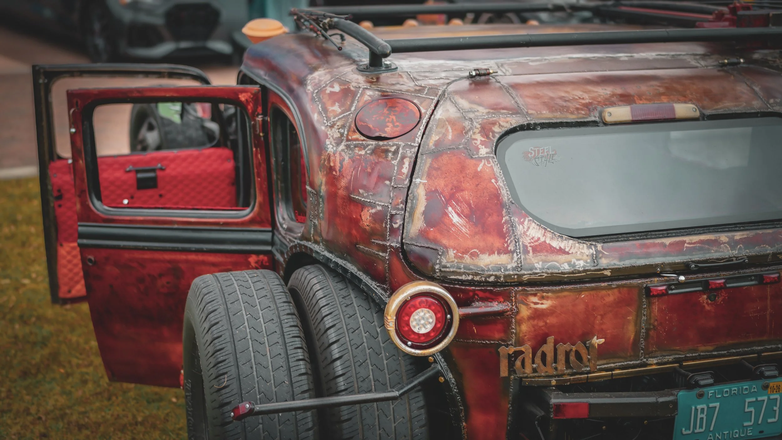A custom vintage vehicle with a distressed red paint job, large rear tires, and an open door showing a quilted red interior. The back has a round taillight and a Florida license plate reading JB7 573, with a rusty, patchwork appearance and a decorati