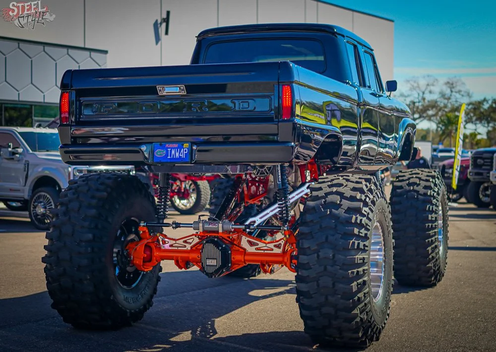 A lifted black Ford pickup truck with oversized off-road tires, altered suspension, and an orange differential, parked outdoors near other vehicles at a car show.