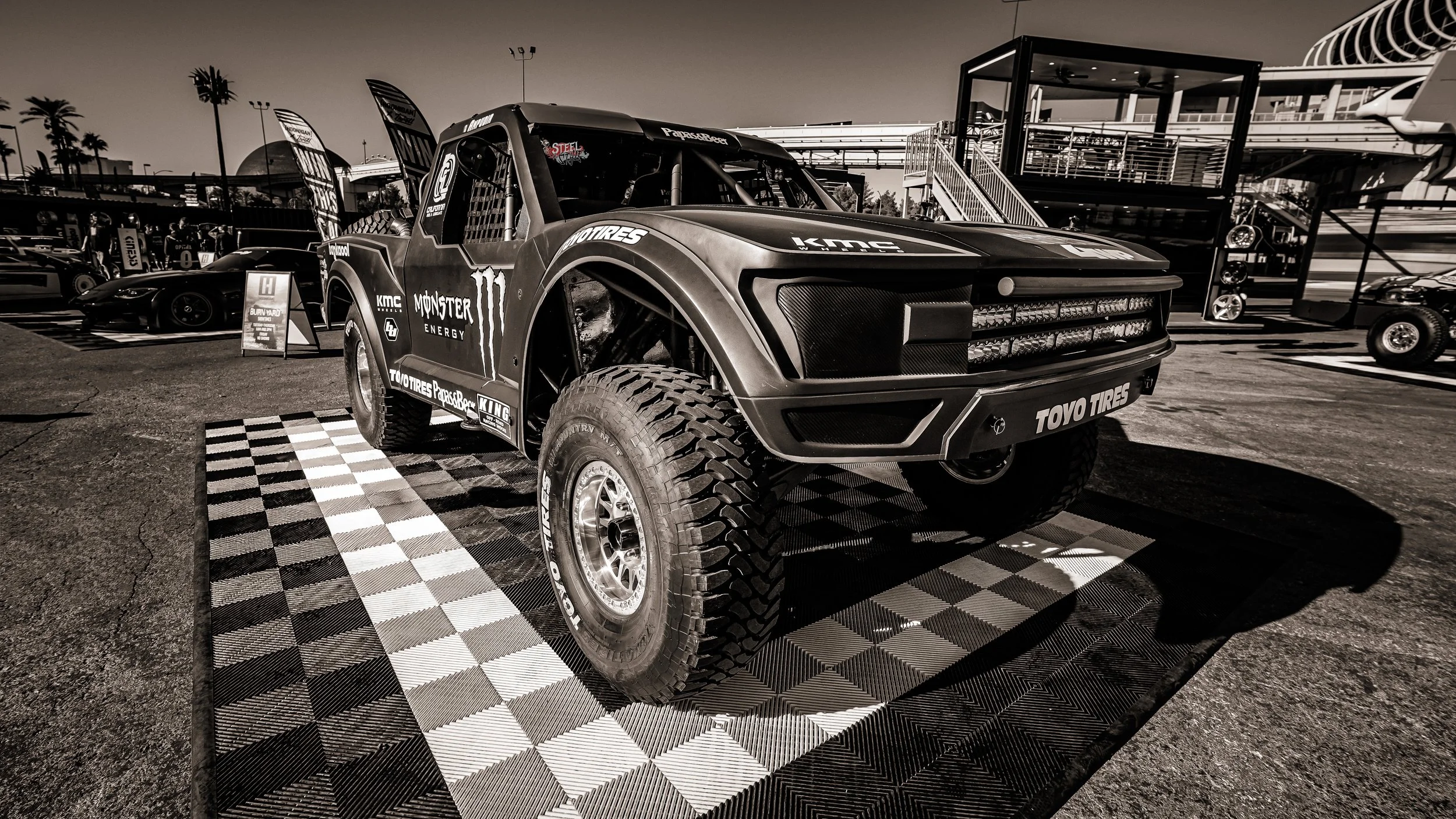 A race truck displayed at a motorsport event, with a checkered pattern floor beneath it, surrounded by other vehicles and structures in the background.
