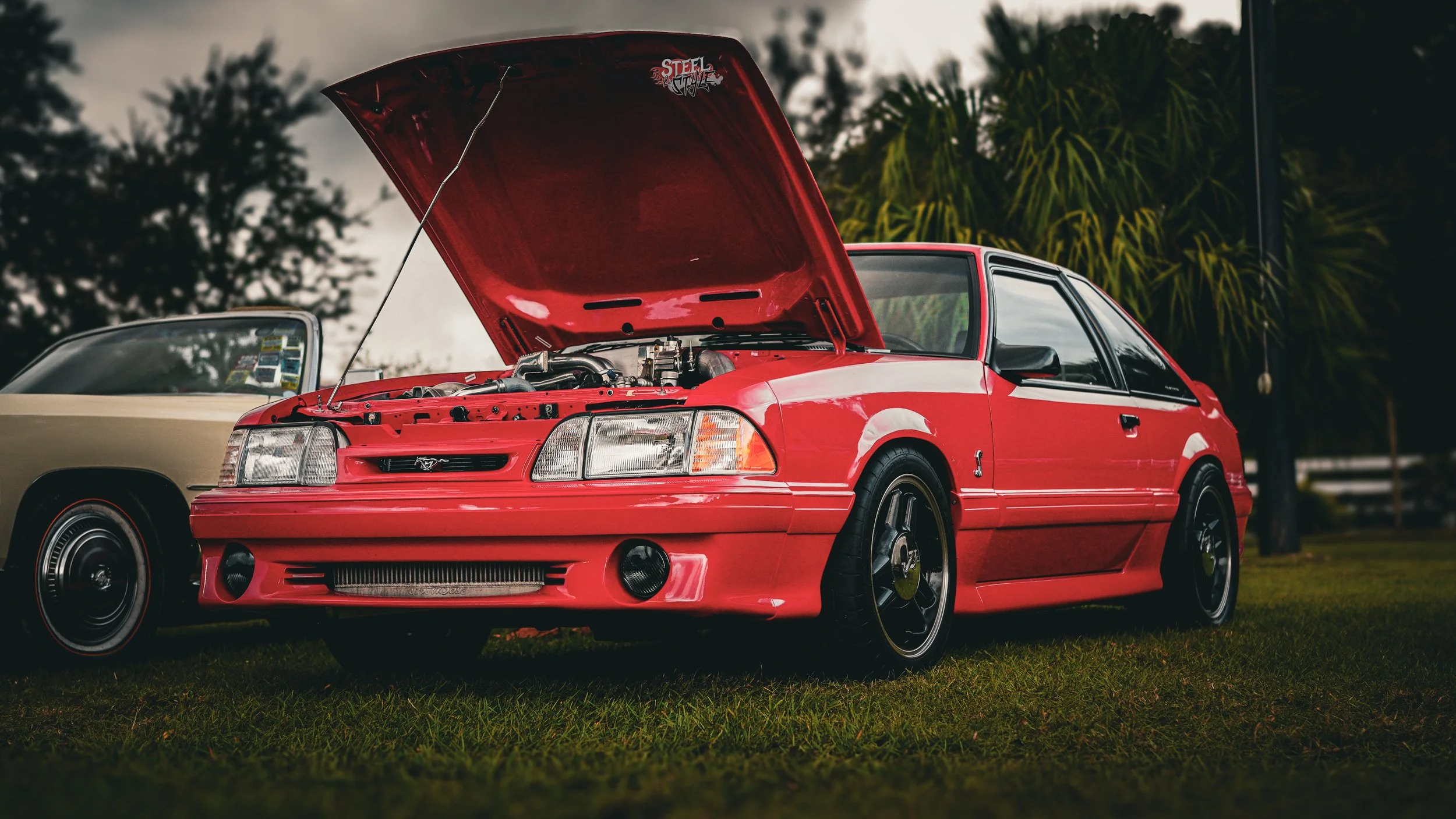 Red Ford Mustang car with open hood parked on grass, with a beige car in the background and trees surrounding.
