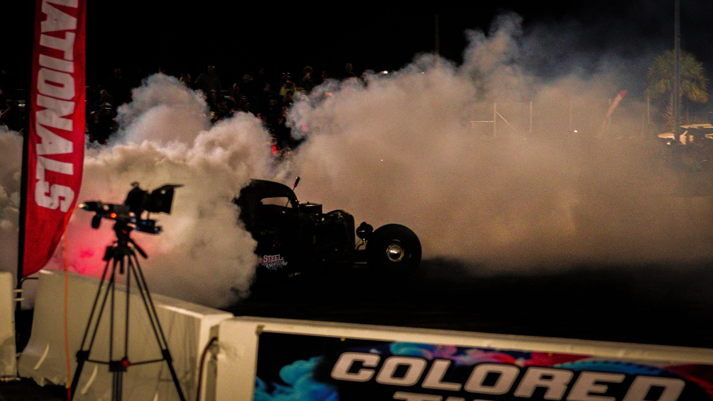 Nighttime drag racing event with a vintage car emitting smoke, surrounded by smoke clouds and spectators. A camera on a tripod captures the scene, and race banners are visible.