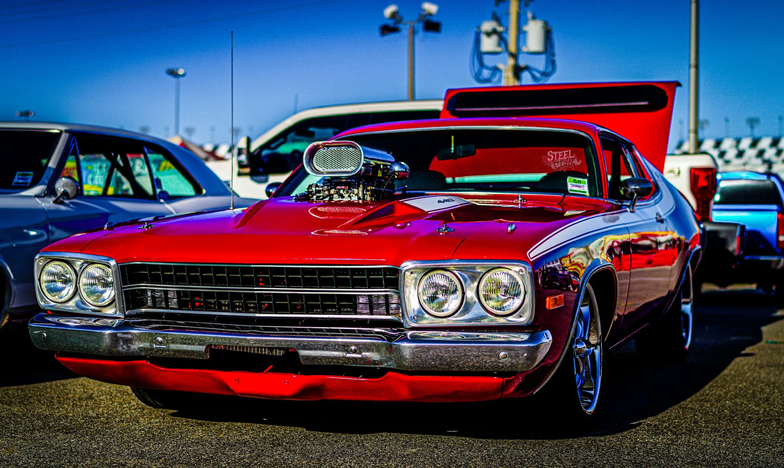 Red vintage muscle car with a supercharger on the hood, parked among other vehicles at a car show.