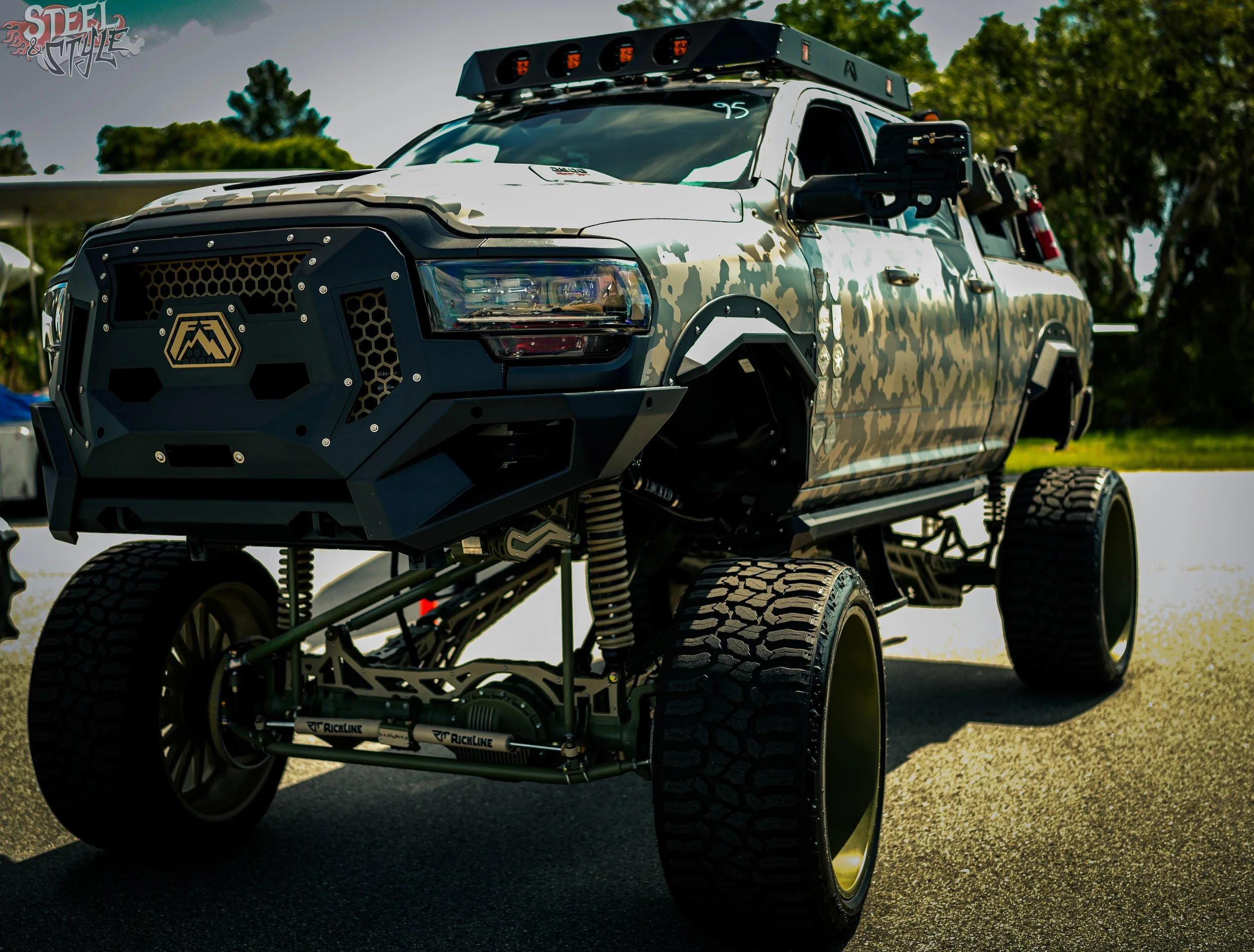 A modified pickup truck with a camouflage paint job, large off-road tires, a custom black grille with a logo, LED light bar on the roof, and added equipment on the sides. The background includes trees and a cloudy sky.