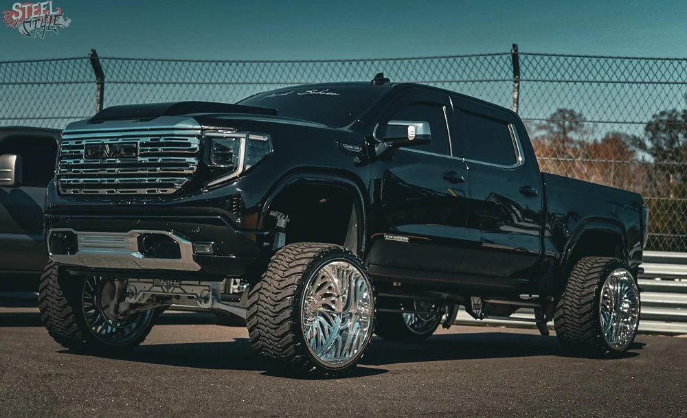 Black lifted pickup truck with oversized shiny chrome wheels parked on a racetrack with a fence and trees in the background.
