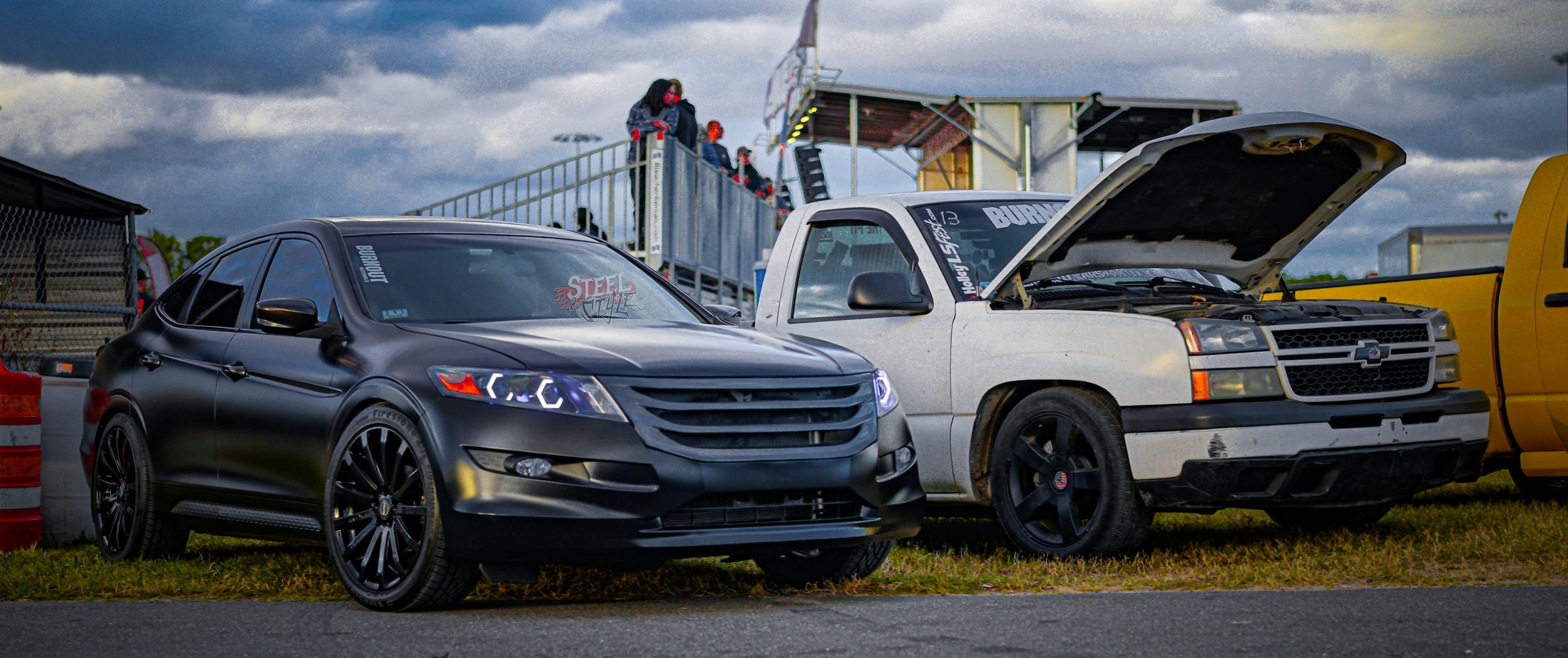 A black modern sedan parked next to a white vintage pickup truck with its hood open, at a race track with spectators in the background.