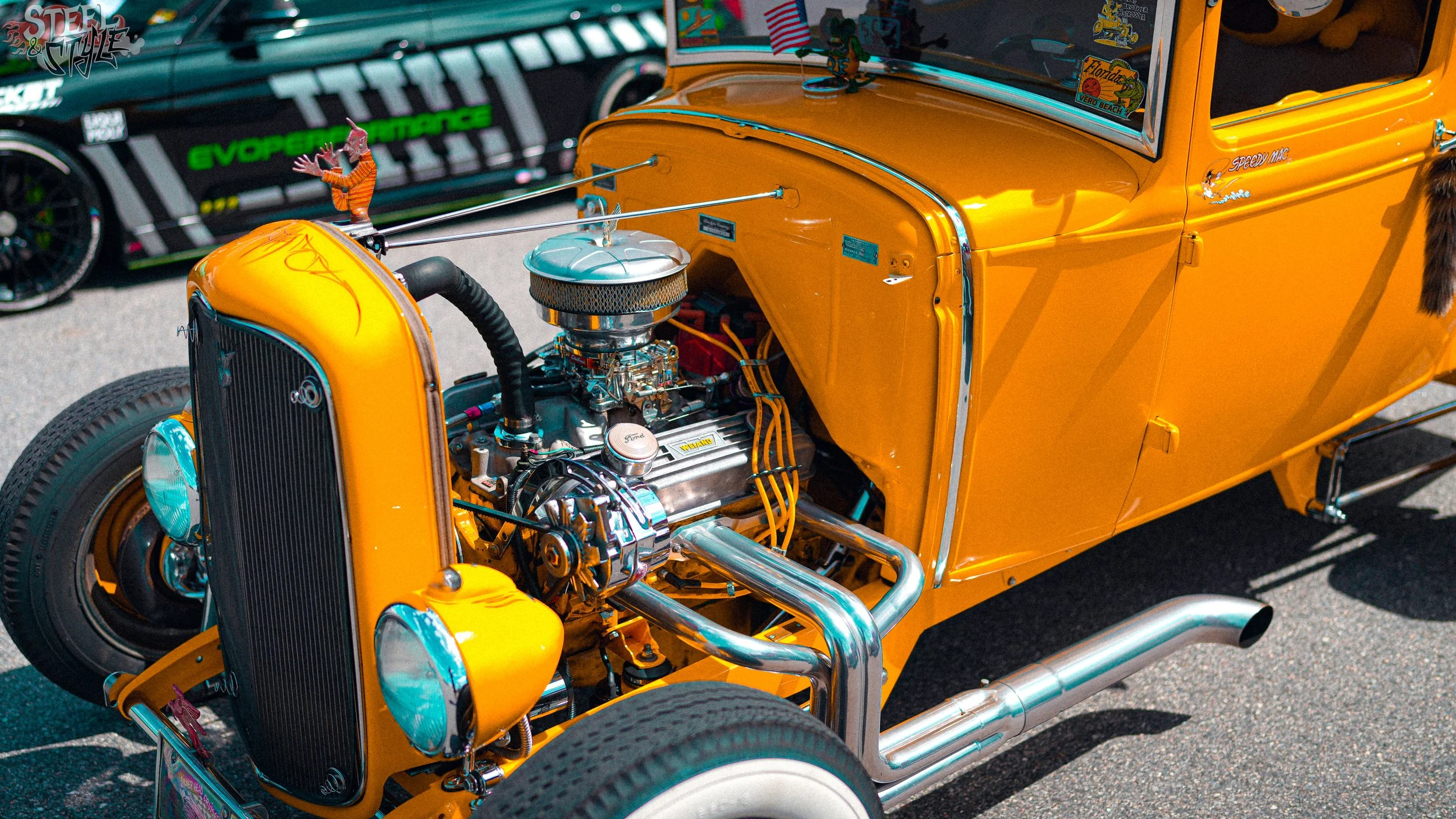Close-up of a vintage yellow hot rod car with an exposed engine, parked at a car show. In the background, there's a race car with colorful decals and graphics.