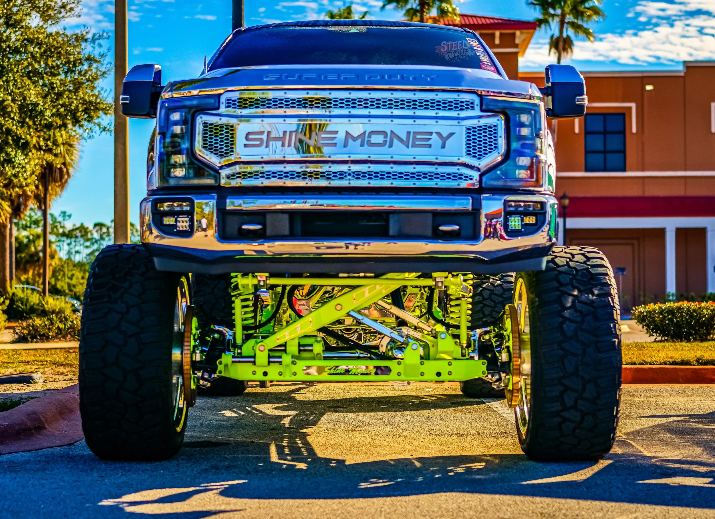 Front view of a lifted custom truck with bright yellow suspension, large tires, and a chrome grille with the words 'Shine Money.' in the windshield reflection.