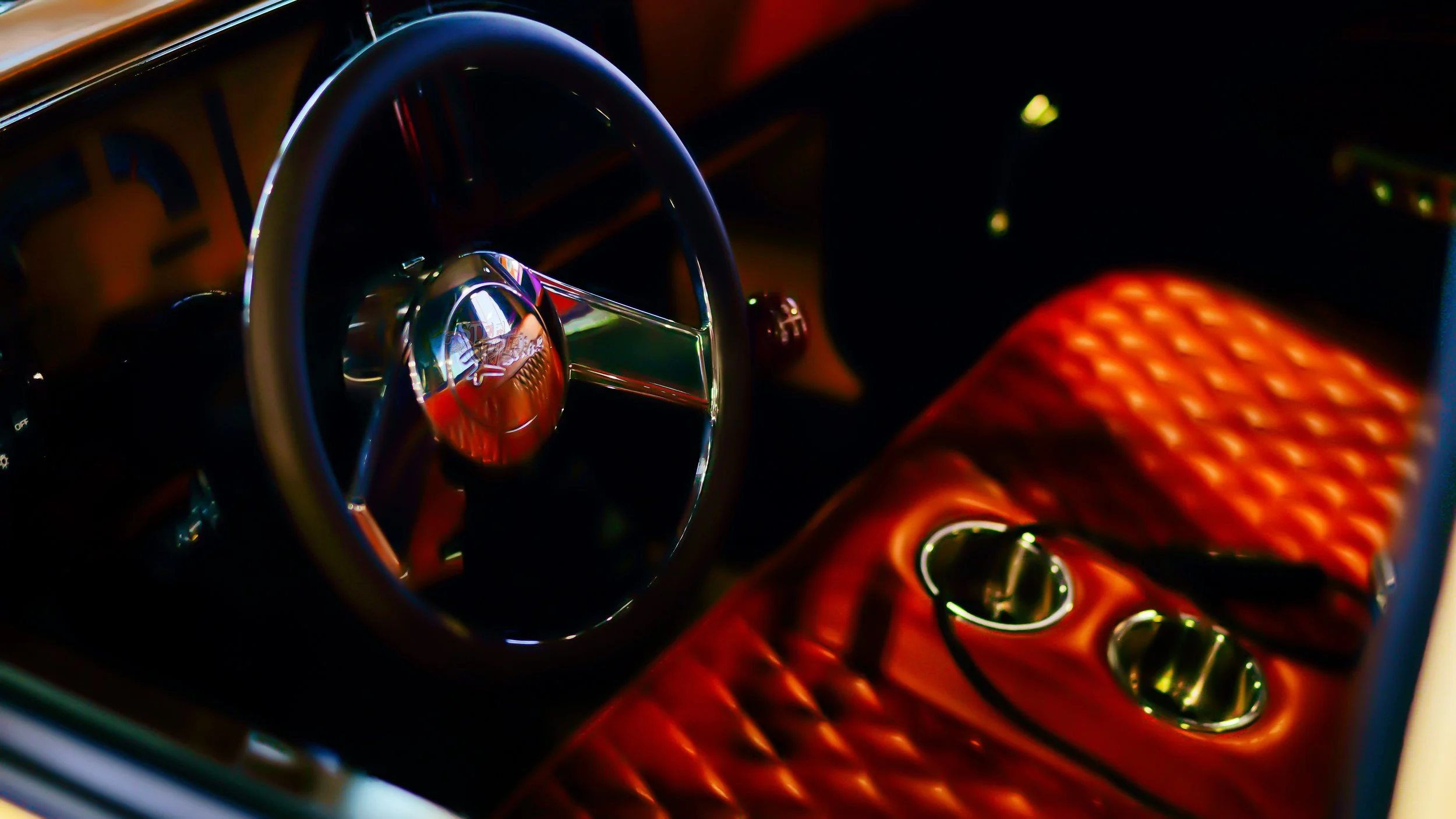 Close-up of a vintage car's interior with a shiny steering wheel and a quilted, orange leather seat with built-in cup holders.