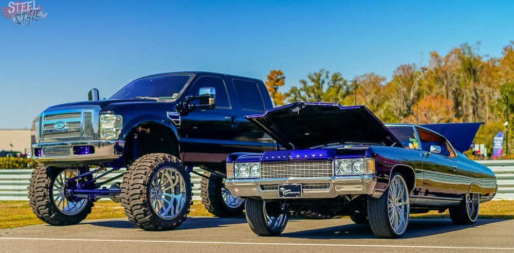 A large black lifted pickup truck with big off-road tires parked next to a classic black Cadillac convertible with its hood open. The scene is outdoors on a sunny day with trees and buildings in the background.