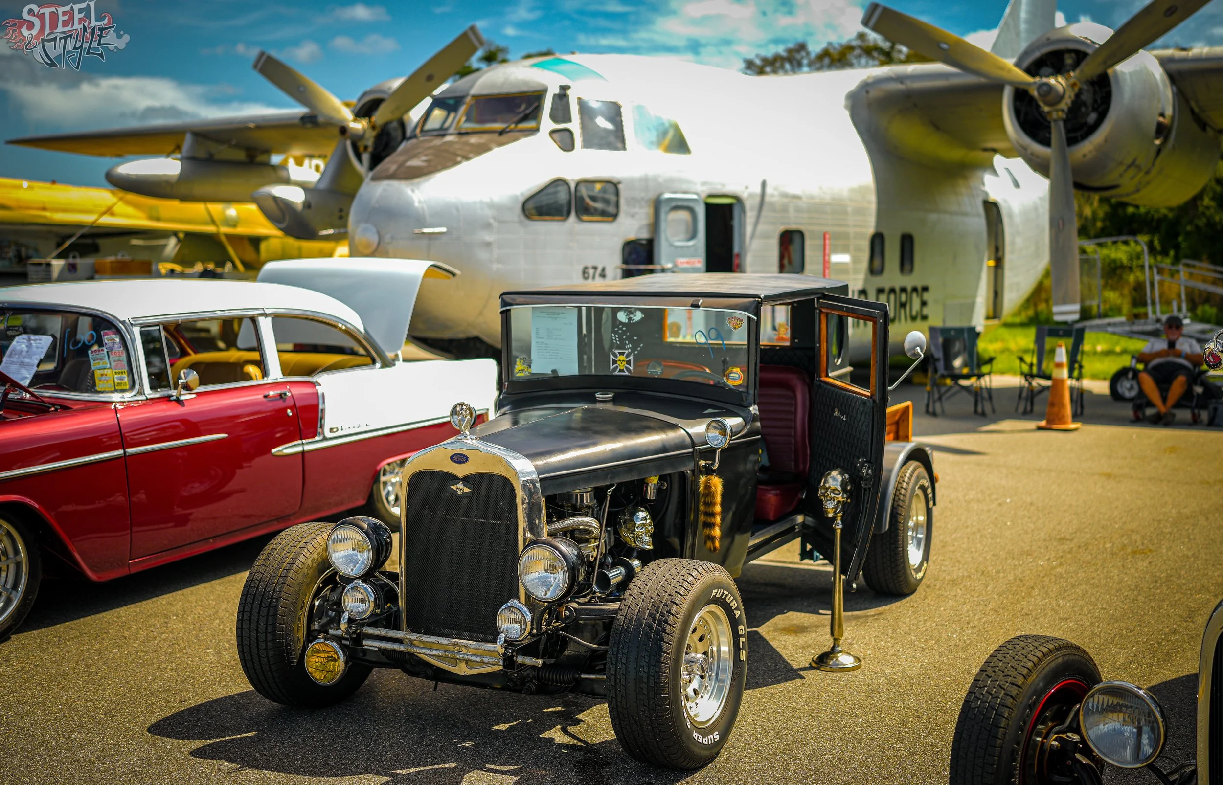 A vintage car and a hot rod car on display at an outdoor car show, with a large military aircraft in the background and a person sitting on a chair.