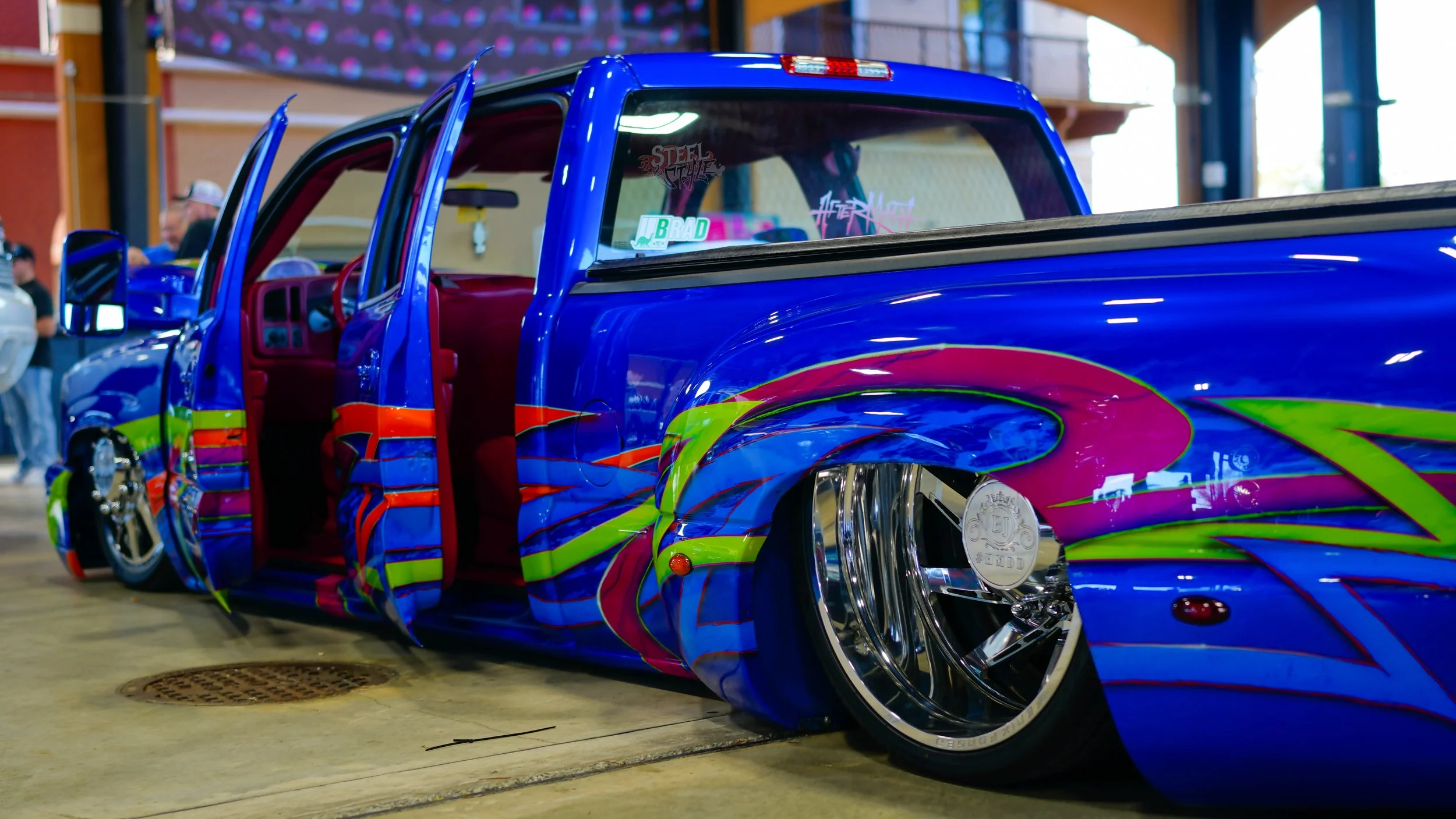 Colorfully customized blue pickup truck with vibrant pink, green, and orange designs, at an indoor car show with open doors and people in the background.