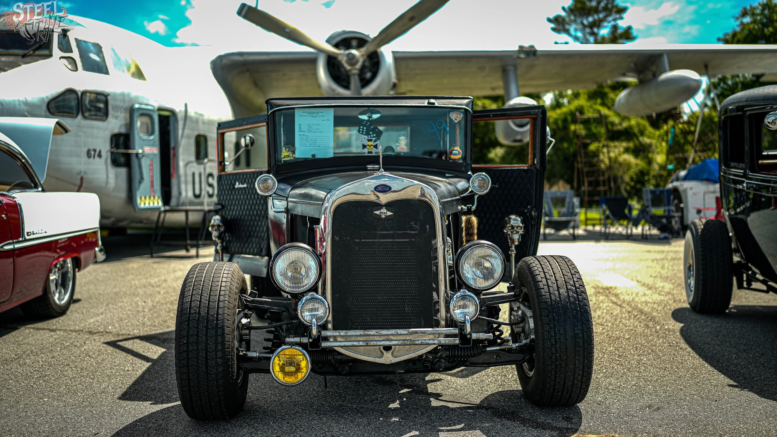 A vintage black and chrome Ford hot rod car parked at an outdoor car show, with a large airplane in the background and other classic cars around.