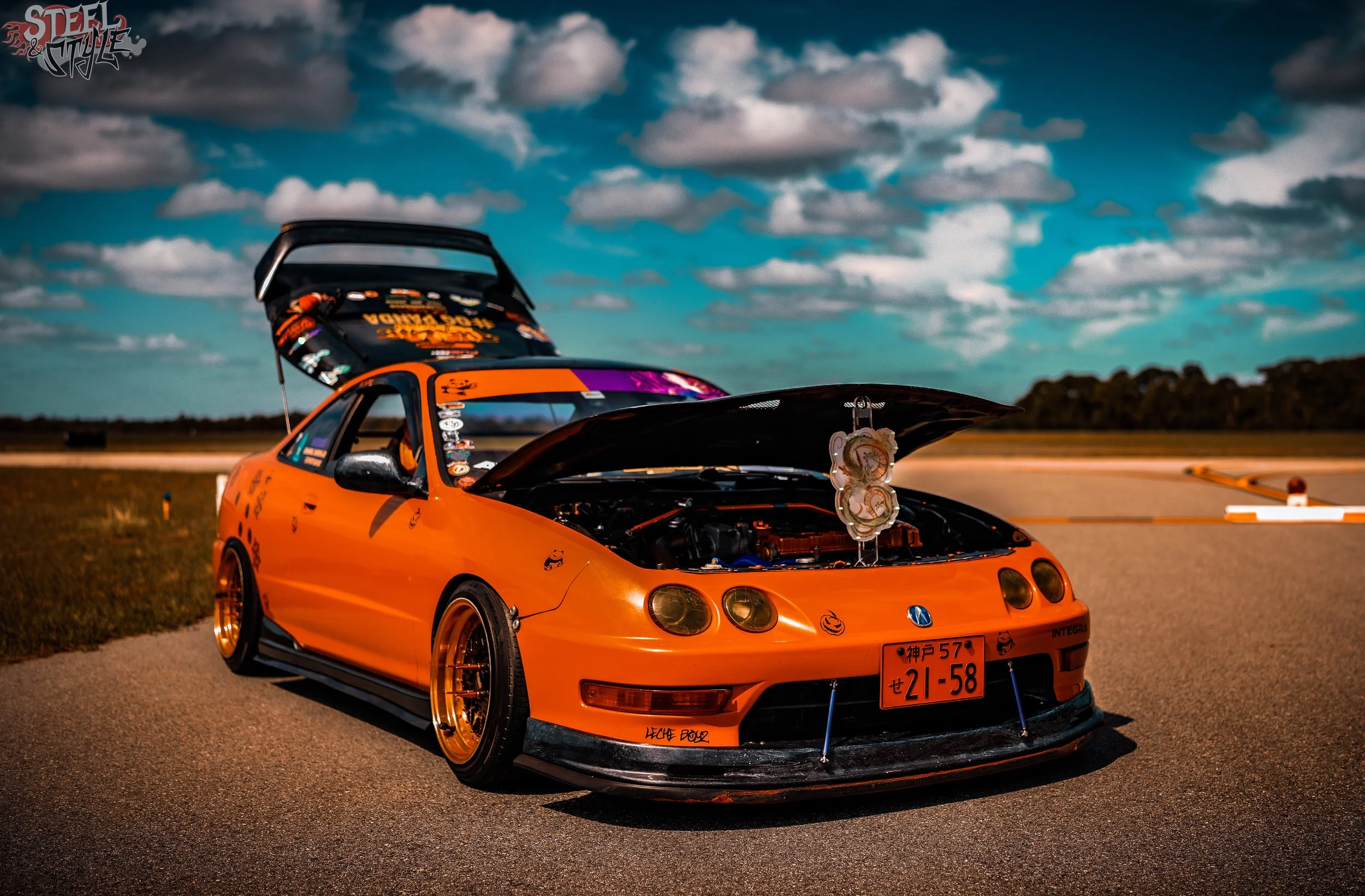 A bright orange sports car with its hood open, parked on an open road or racetrack under a cloudy sky, with decals and modifications visible.