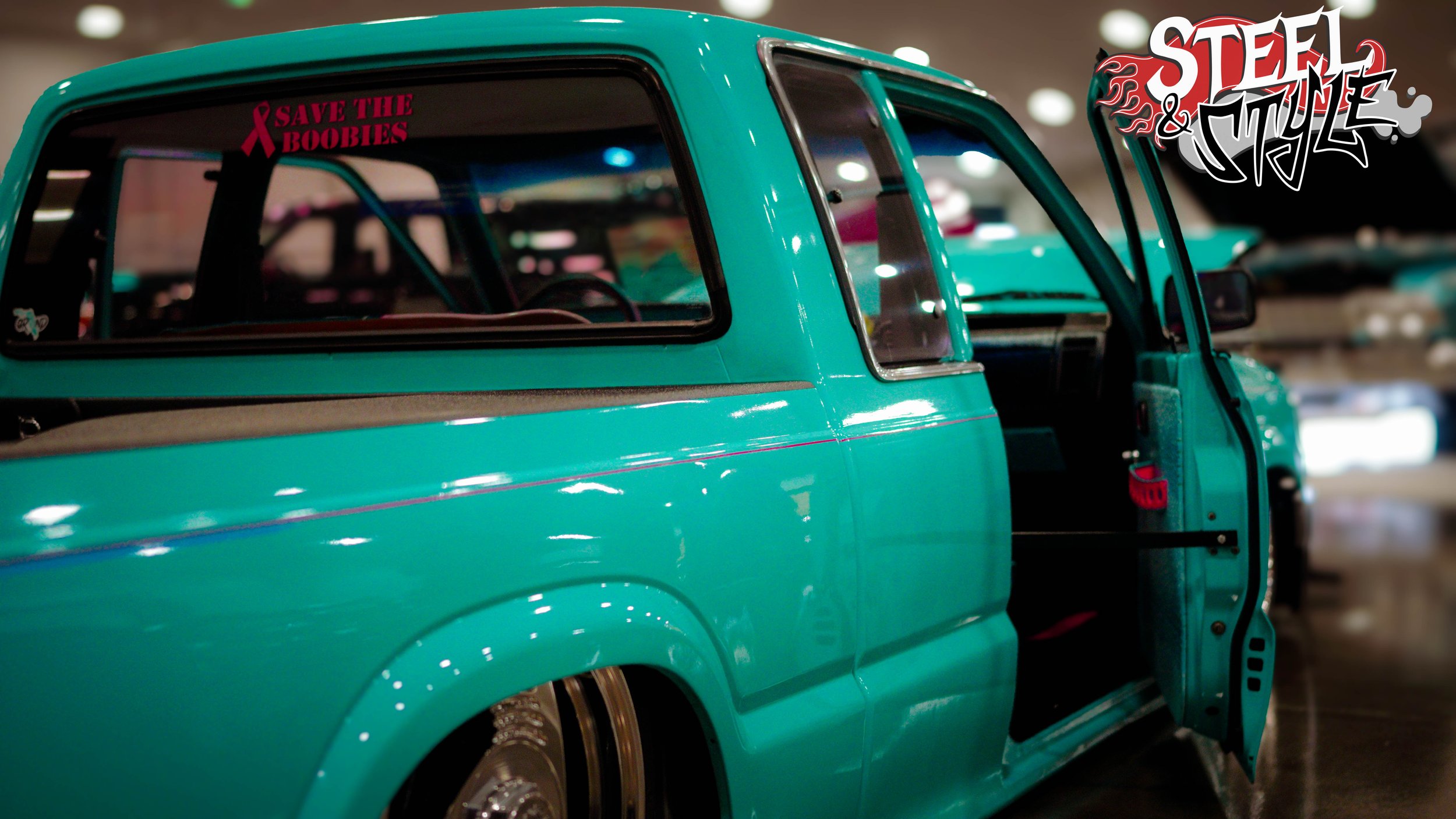 A teal vintage truck displayed indoors with an open passenger door. There are stickers on the window, including one that says 'Save the Boobies' with a pink ribbon and another with a stylized logo reading 'Steel & Style.'