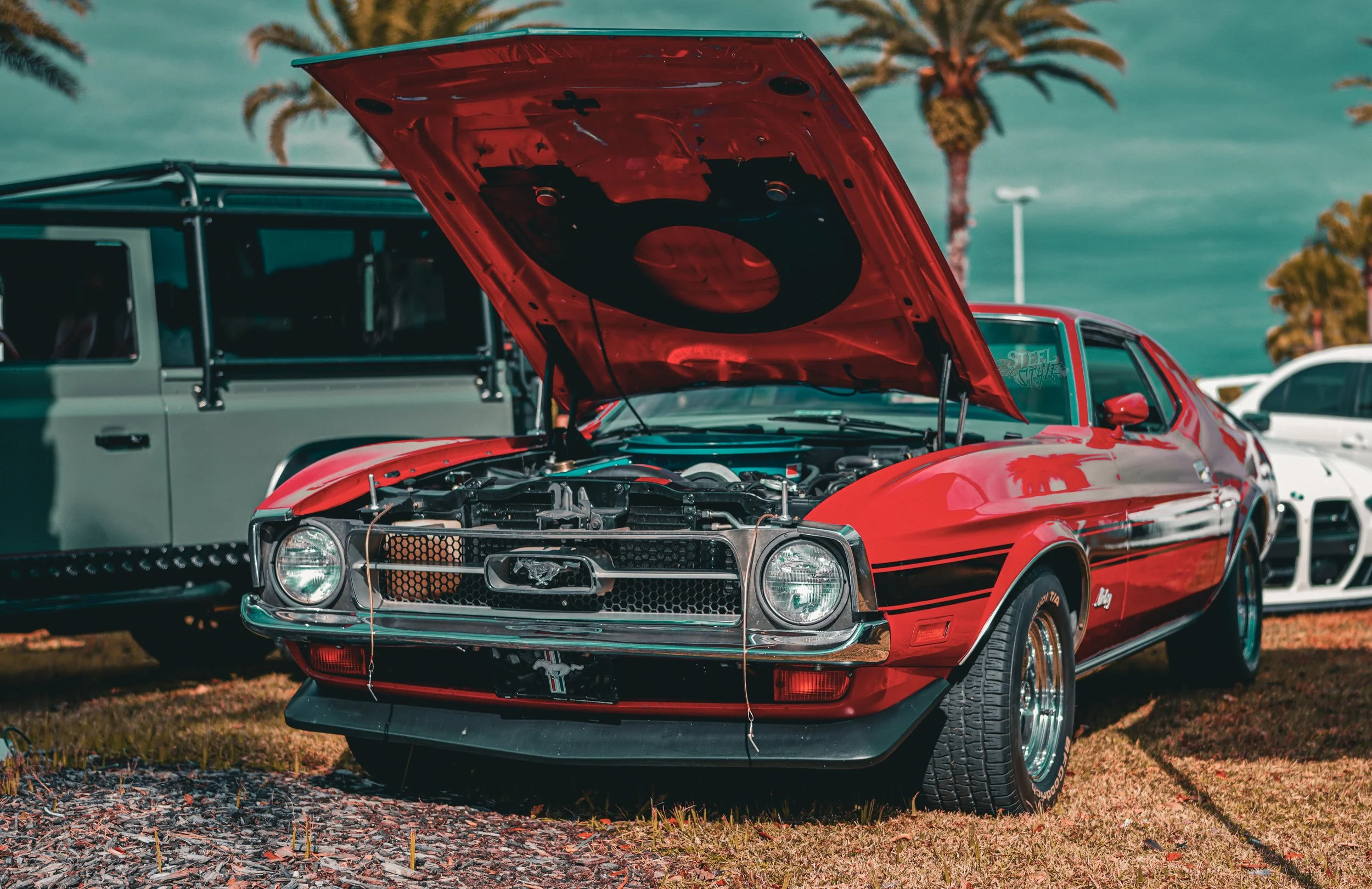 A red vintage Ford Mustang with its hood open at a car show, showcasing its engine. Other cars and palm trees are visible in the background.