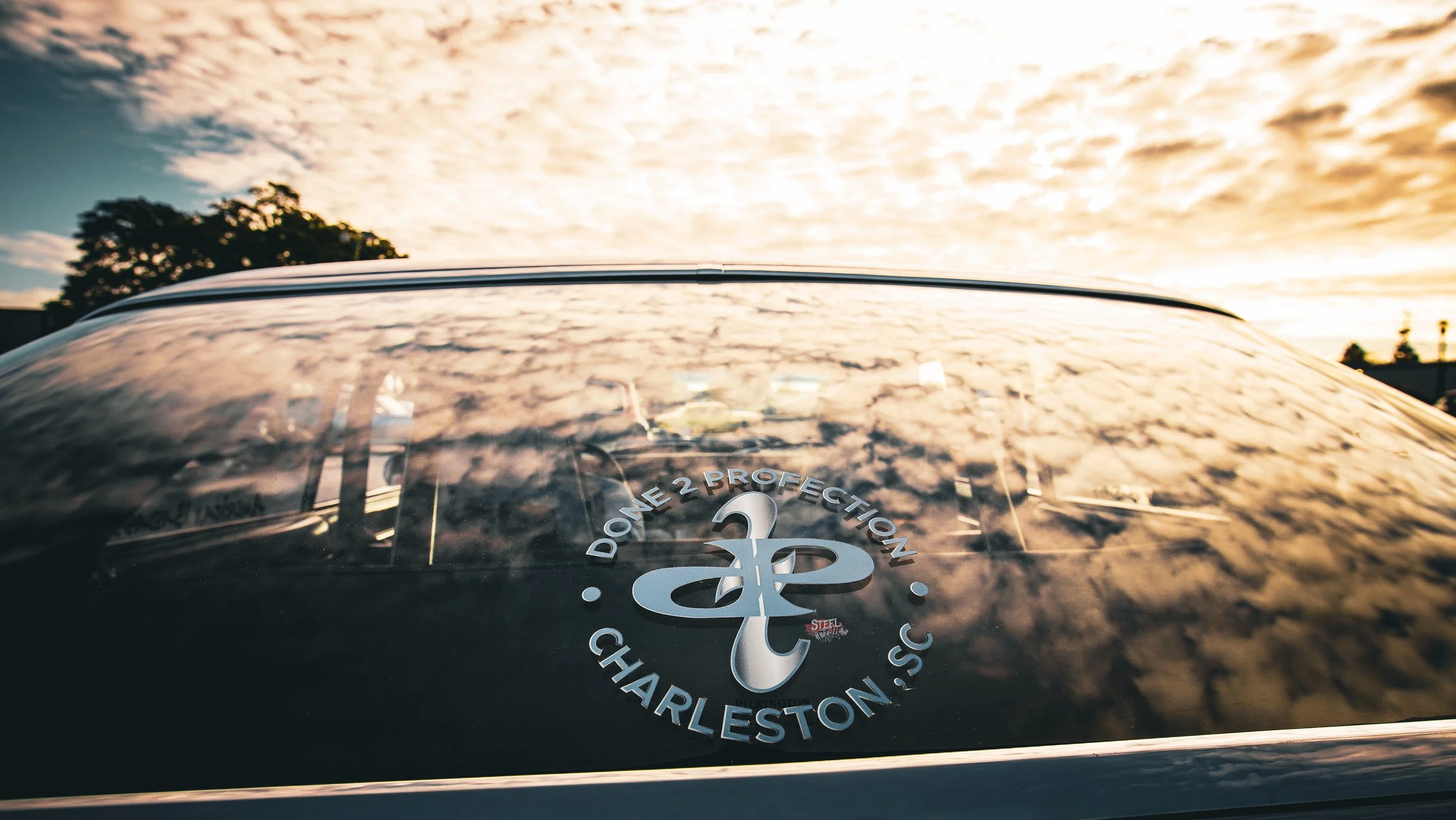 Close-up of a car windshield reflecting a cloudy sky during sunset with a sticker that says 'Done 2 Protection Charleston, SC' and features a stylized whale logo.