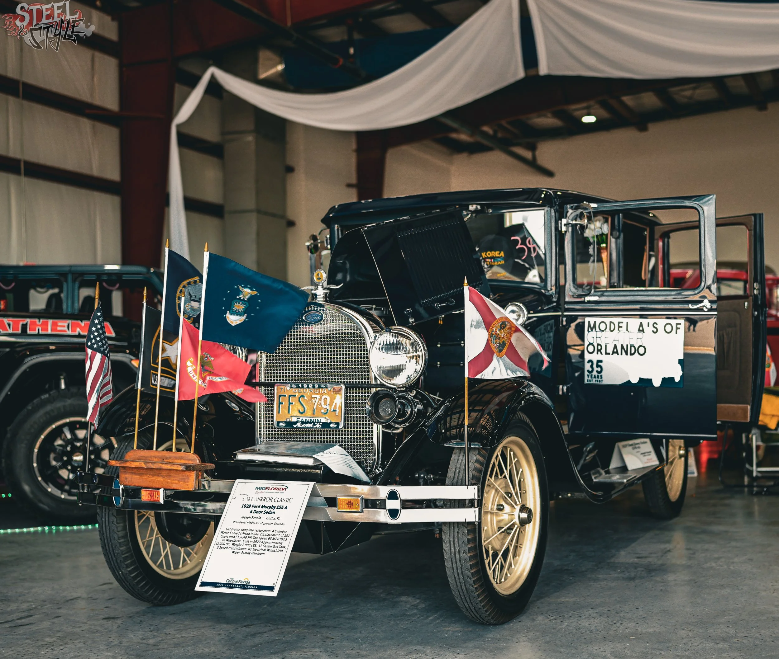 Vintage black car on display at an indoor car show, decorated with flags and a sign with details about the car.