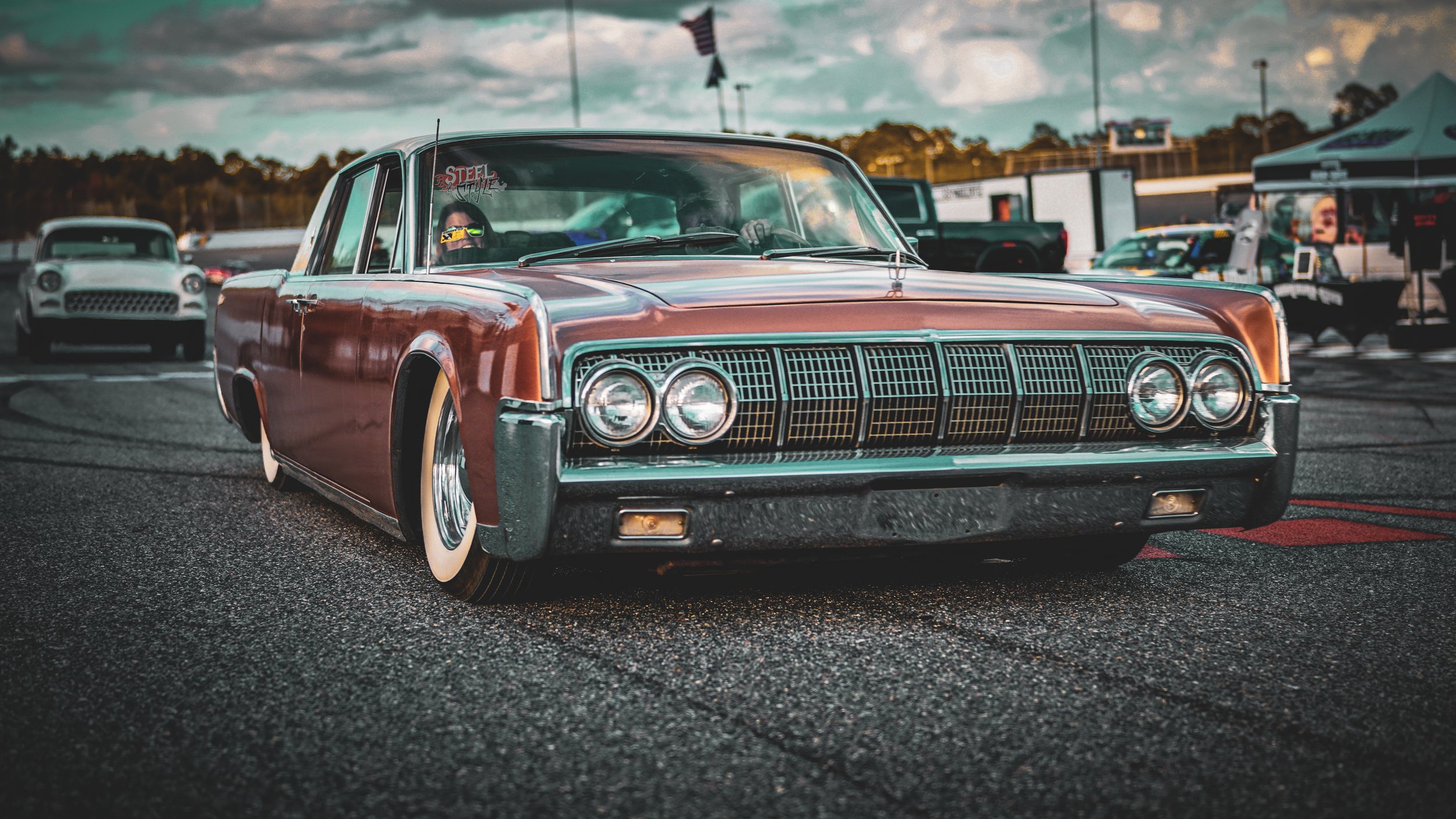 Vintage red car with whitewall tires parked on a racetrack during an event, with other cars and tents in the background.