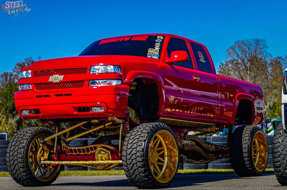 A red Chevrolet pickup truck modified with large gold wheels and lifted suspension, displayed outdoors with a clear blue sky and trees in the background.