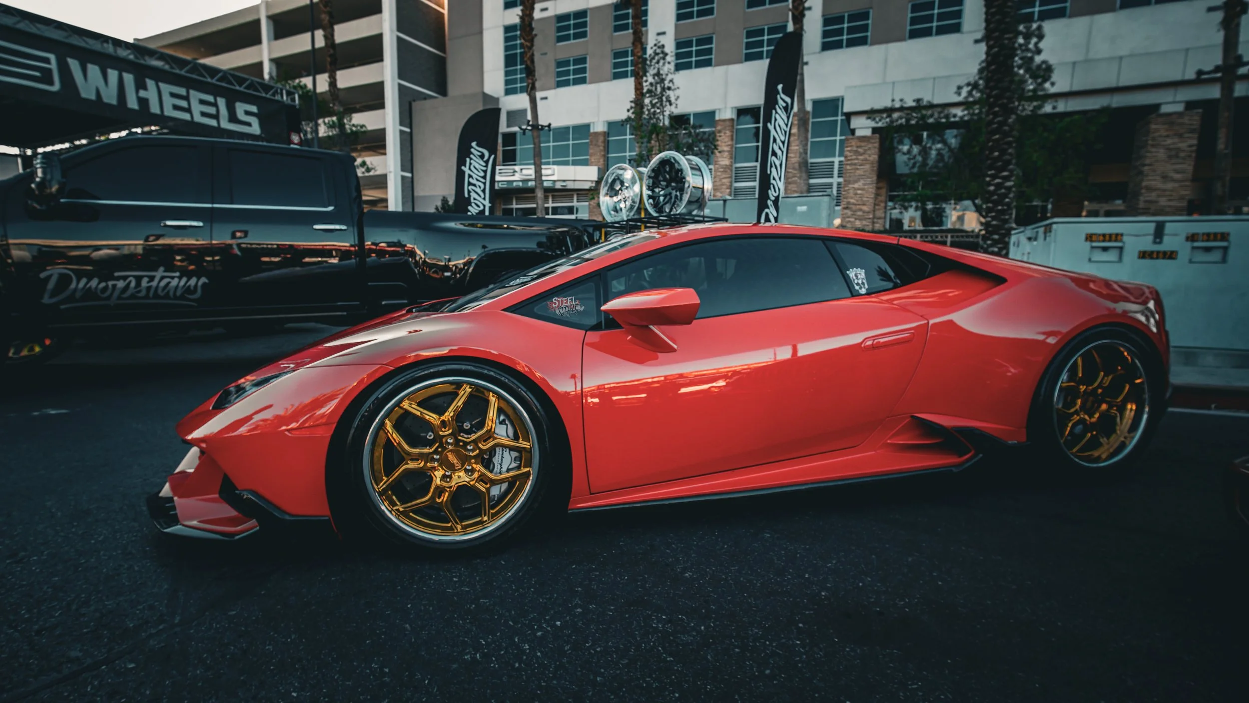 A red Lamborghini sports car with gold wheels parked on a street during an outdoor car show.