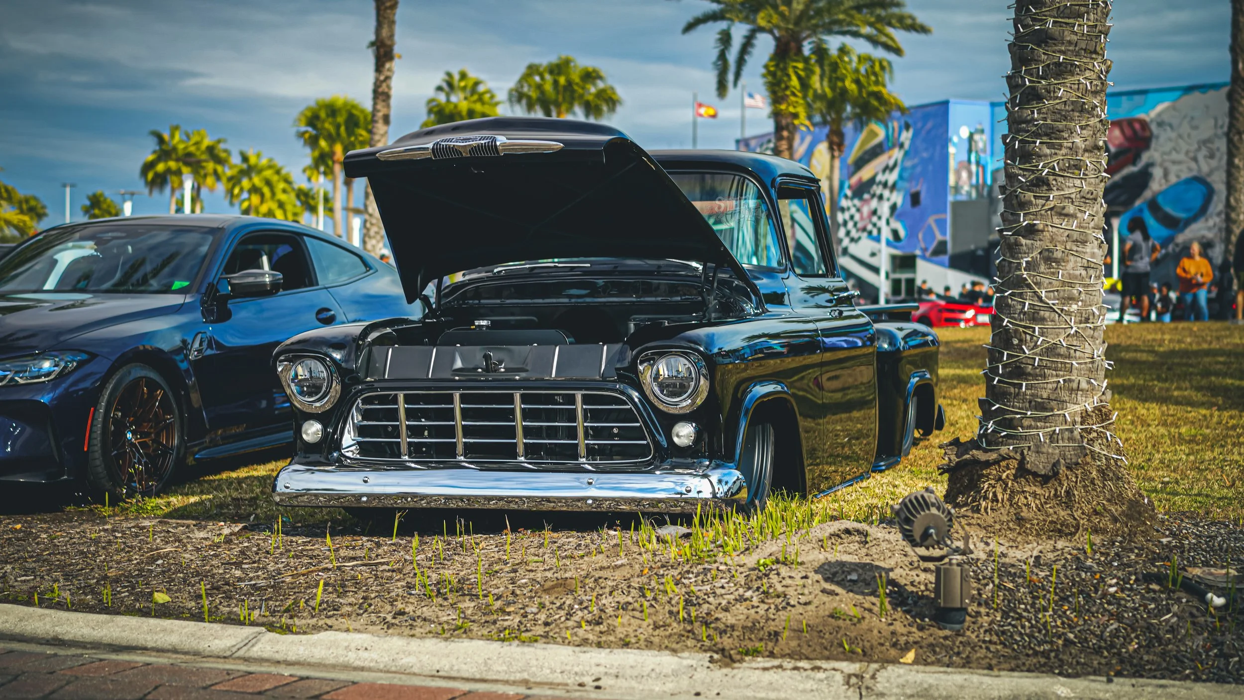 Black vintage pickup truck with open hood parked on grass at outdoor car show, next to a blue modern sports car, palm trees, colorful murals, and people in the background.