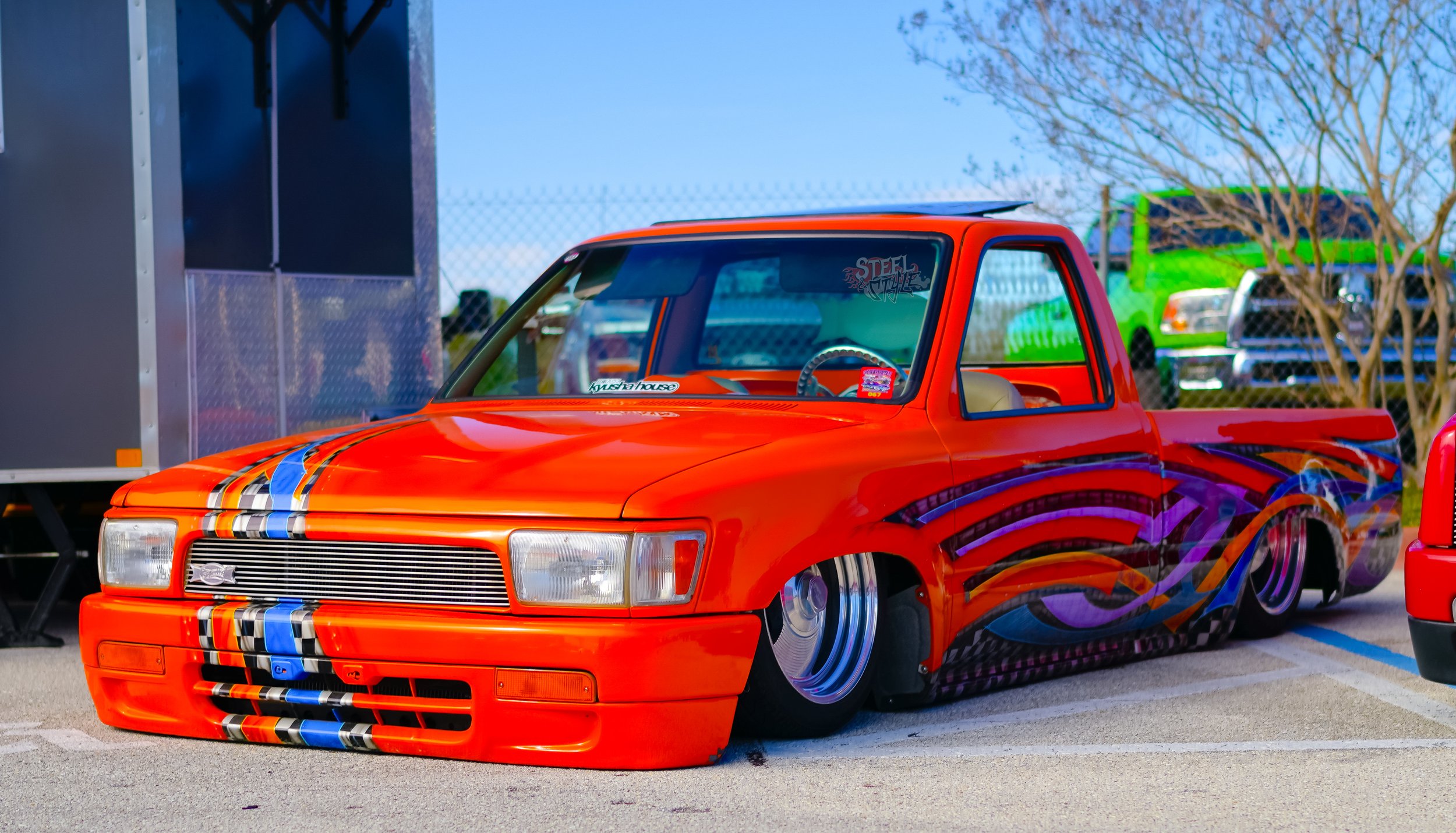 A customized red pickup truck with low ride height, featuring blue, purple, and orange flame-like graphics on its sides and hood, parked in a lot with other vehicles in the background.