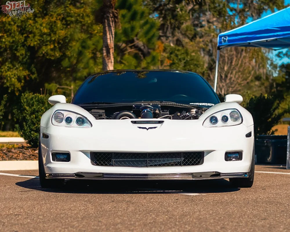 White Chevrolet Corvette sports car with hood open, parked outdoors near trees and a blue canopy.
