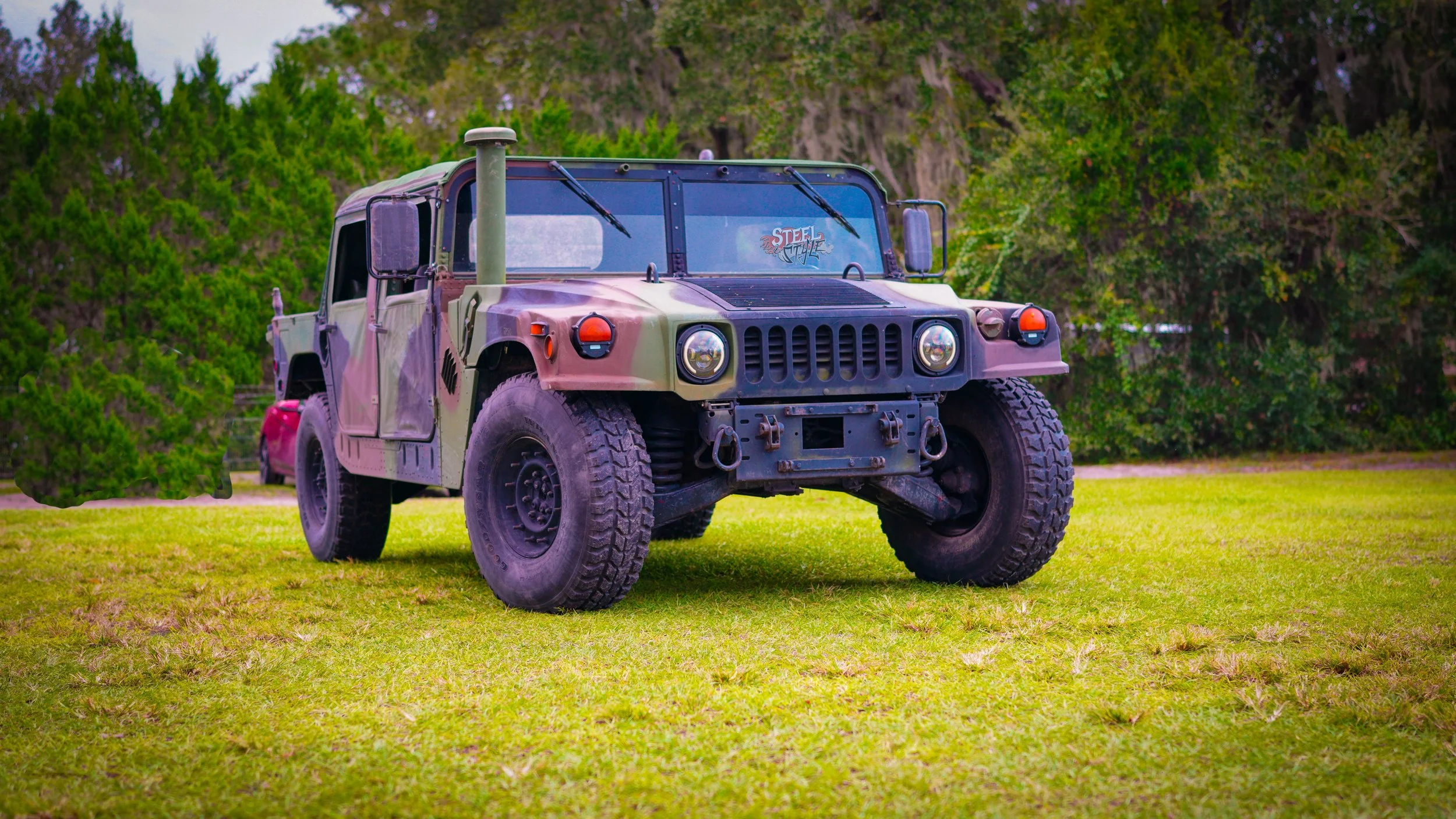 A military-style camouflage vehicle parked on a grassy field with trees in the background.