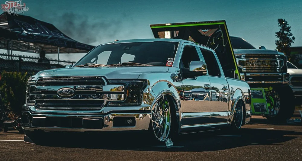 A reflective chrome Ford truck with custom wheels and an open hood, parked at an outdoor car show with tents and other vehicles in the background.