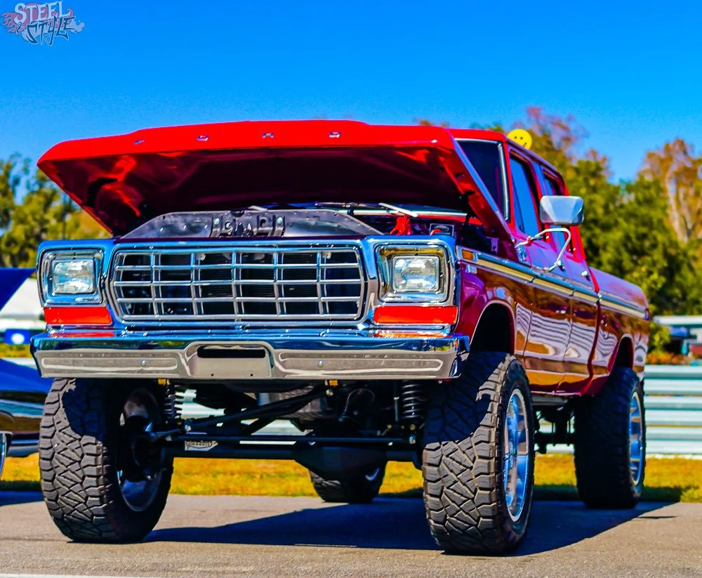 A lifted red and purple vintage pickup truck with its hood open, displayed outdoors during daytime with clear blue skies and trees in the background.