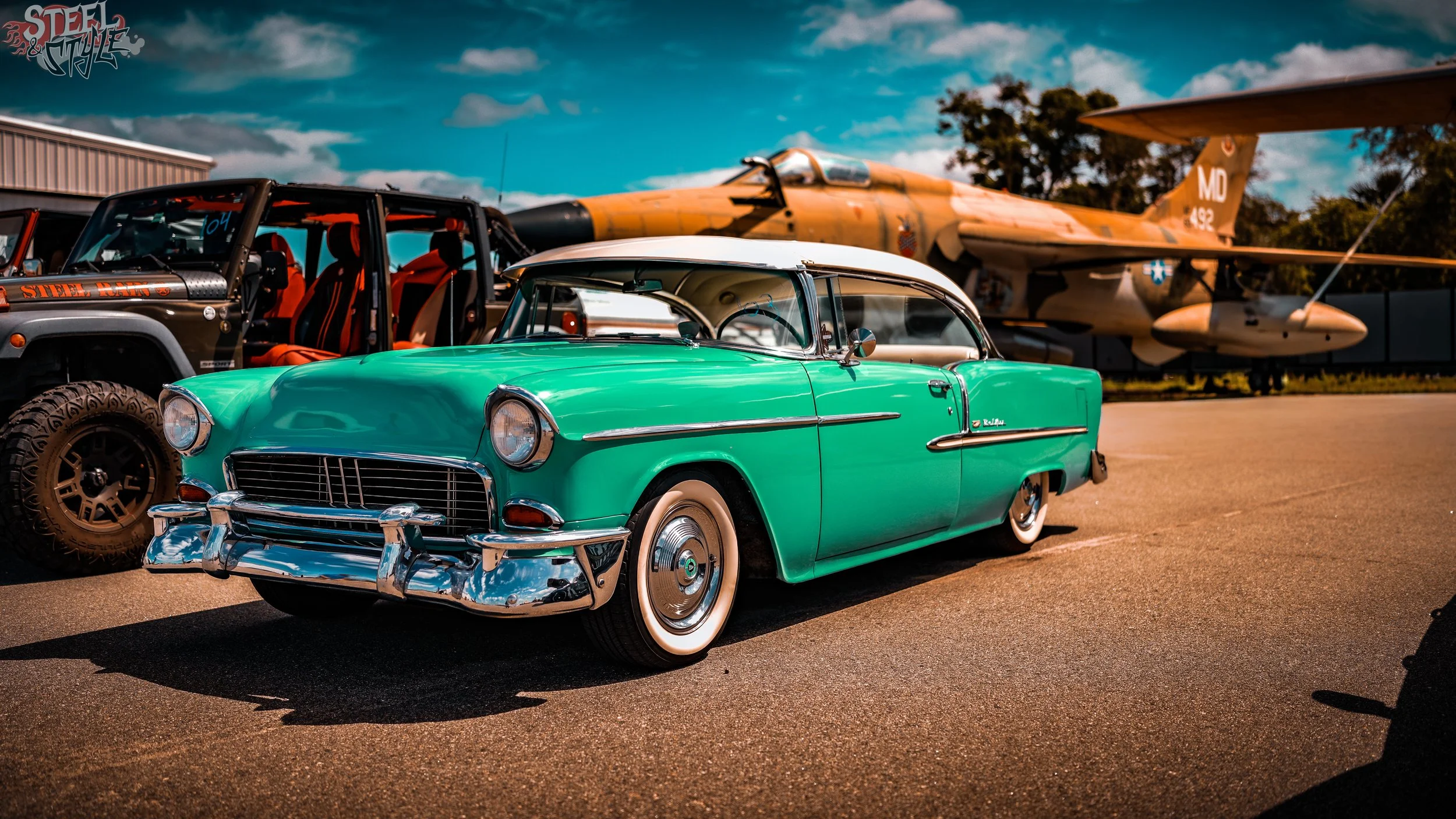 A vintage green and white classic car parked outdoors, with a military jet aircraft and another off-road vehicle in the background under a partly cloudy sky.