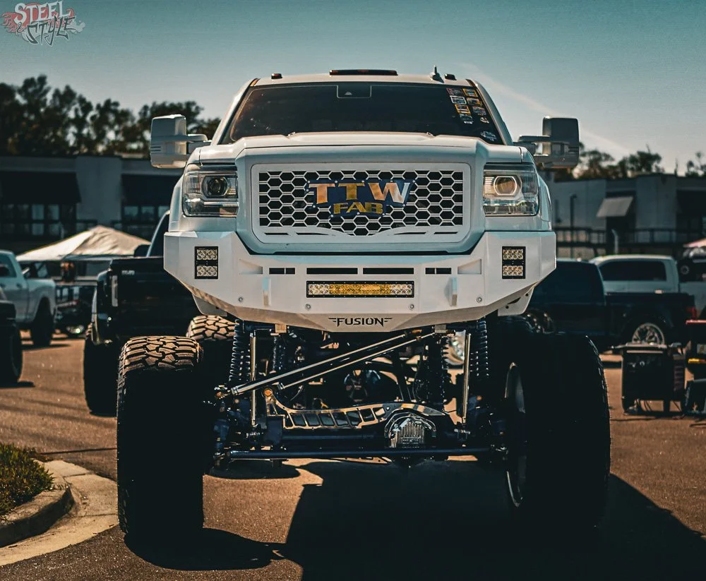 Front view of a lifted white truck parked in a lot, with other trucks and buildings in the background.
