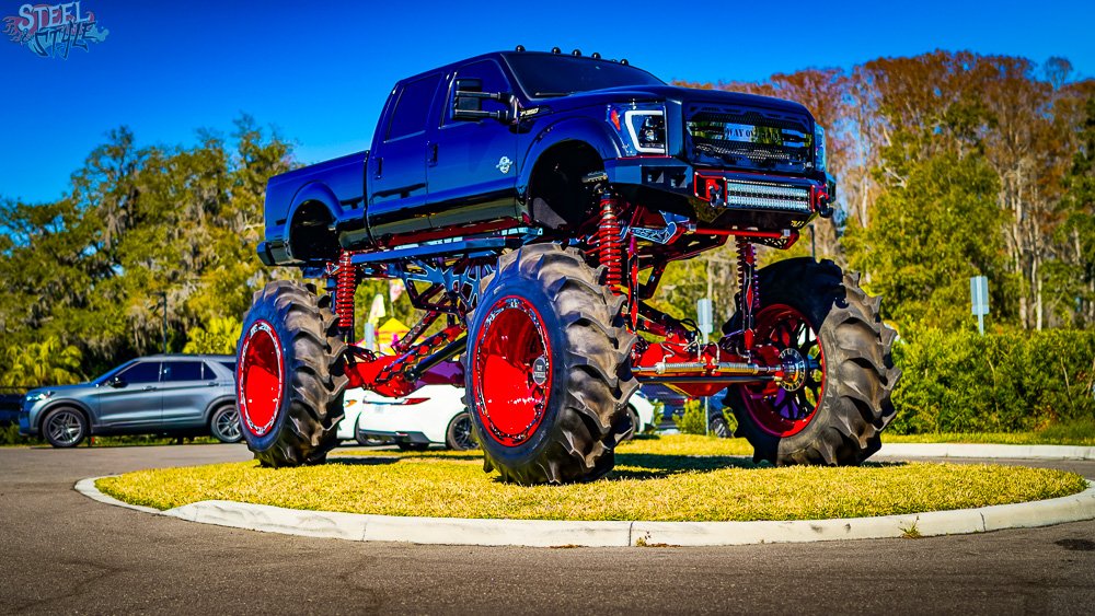 A large monster truck with a black pickup truck body and massive red wheels is on display on a grassy roundabout with cars in the background and clear blue sky.