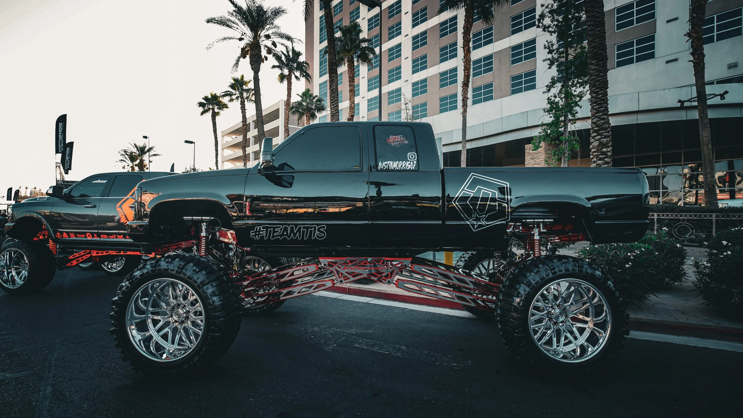 A custom lifted pickup truck with large wheels and tires, parked in front of palm trees and modern buildings.