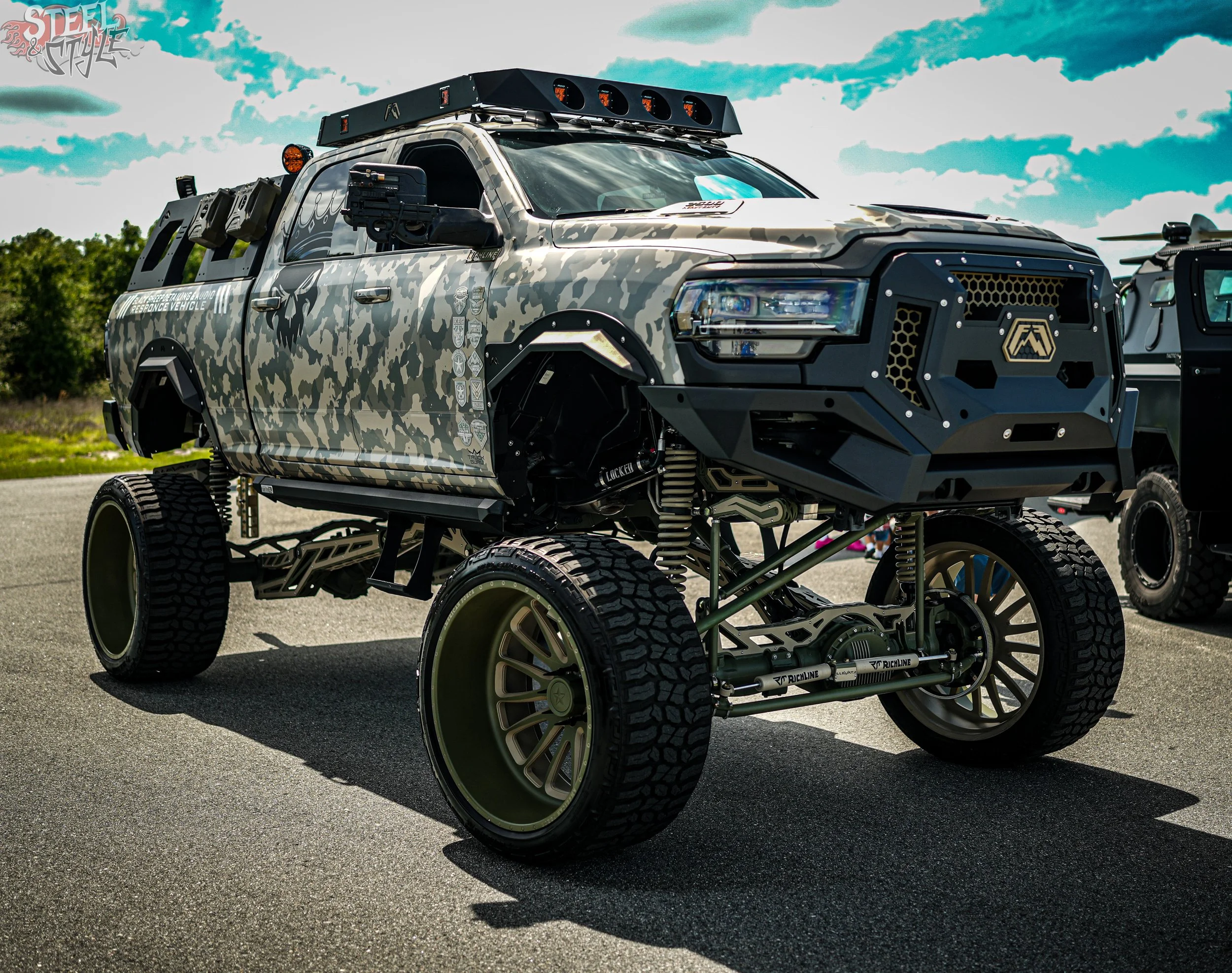 A modified off-road pickup truck with a custom camouflage paint job, large wheels, and a front grille guard, parked outdoors under a partly cloudy sky.