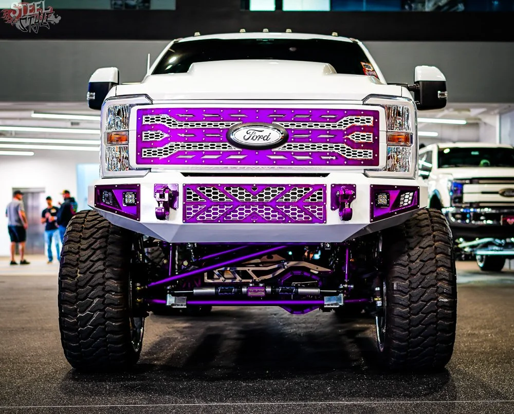 Front view of a modified white Ford truck with purple accents, large off-road tires, and custom grille and bumper at an indoor car show.