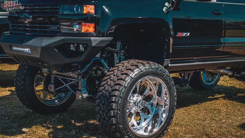 Close-up of a black Chevrolet truck with large off-road tires and custom chrome wheels, displayed outdoors on grass.